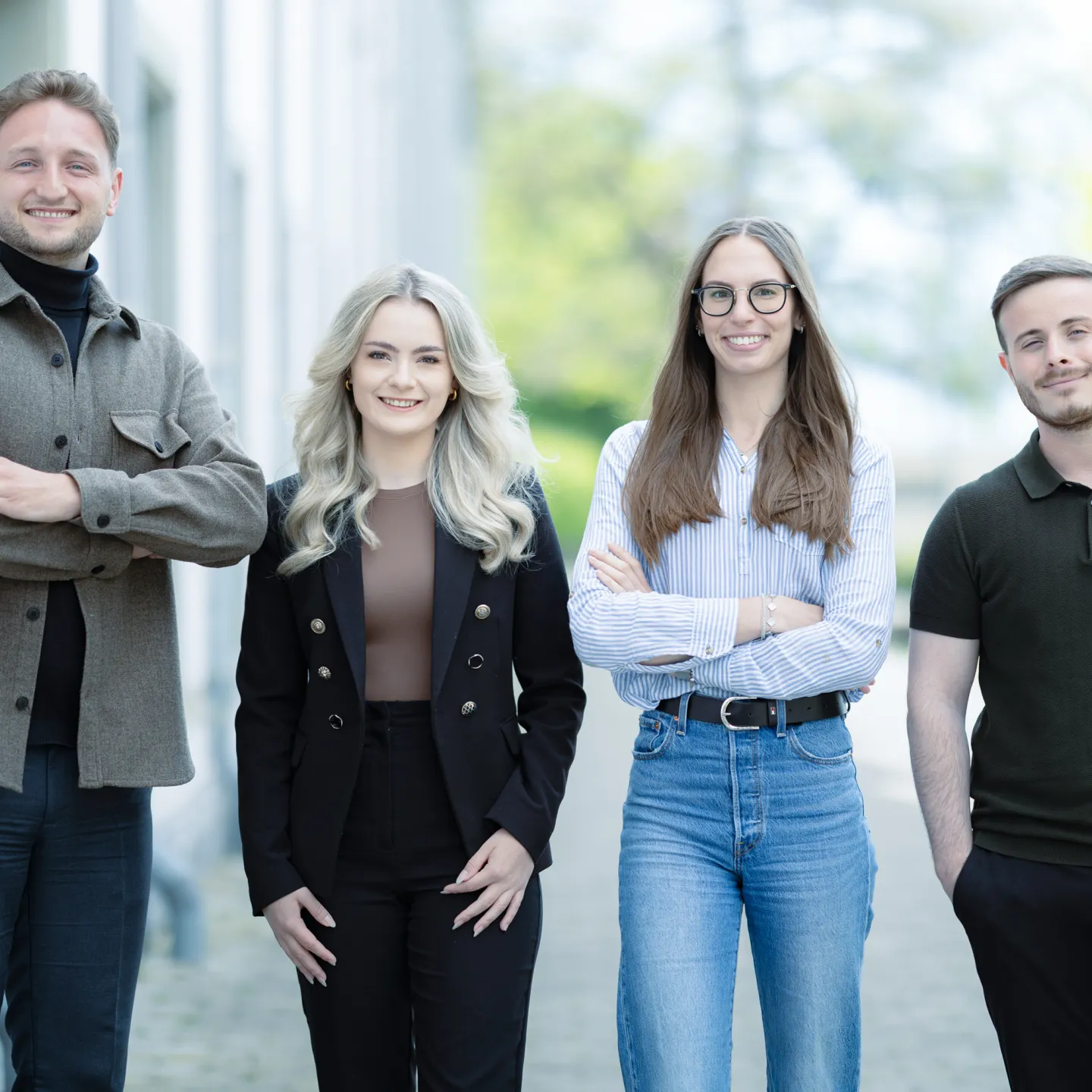 Vier Studierende im Masterstudiengang Information Systems der Universität Liechtenstein stehen nebeneinander auf einem Gehweg vor einem Universitätsgebäude und lächeln in die Kamera. Die Kleidung reicht von Business- bis Casual-Stil.