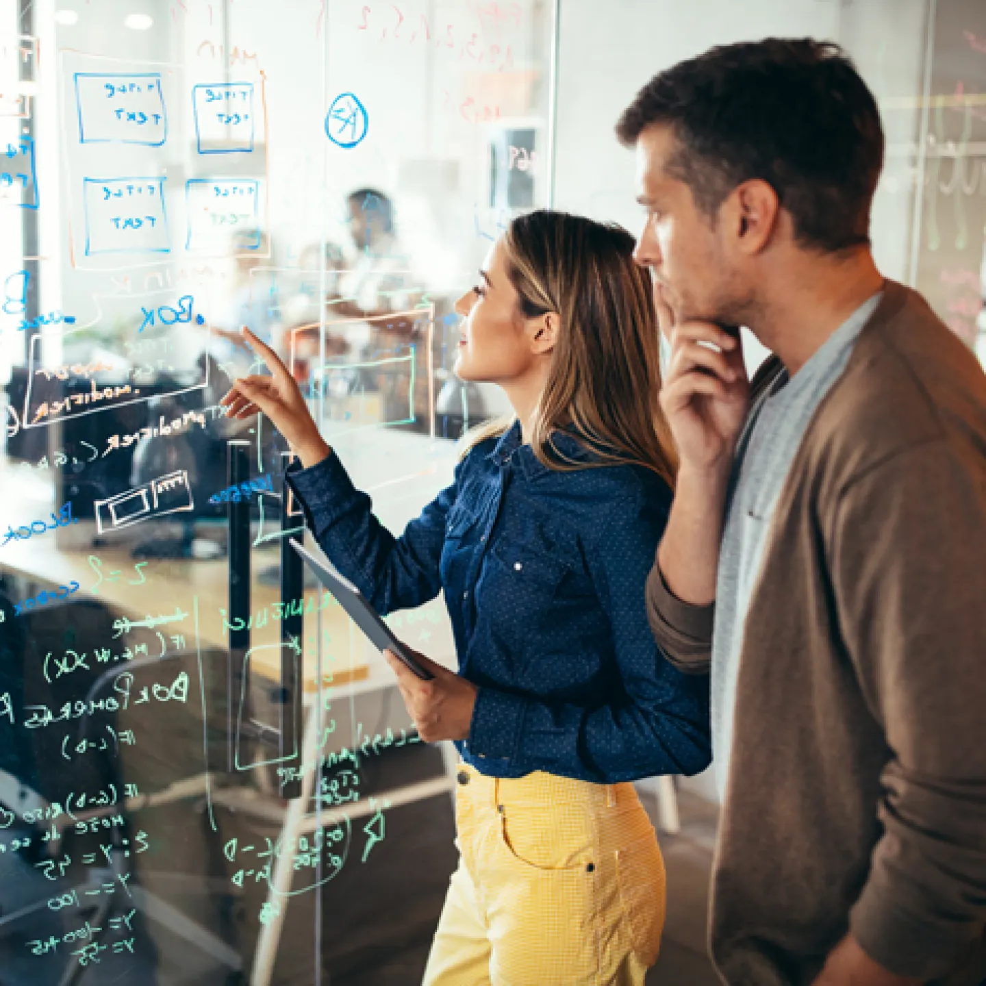 Ein Mann und eine Frau betrachten gemeinsam eine große Glaswand, die mit Notizen und Diagrammen beschrieben ist. Beide wirken nachdenklich und konzentriert, während sie an einer komplexen Aufgabe arbeiten.