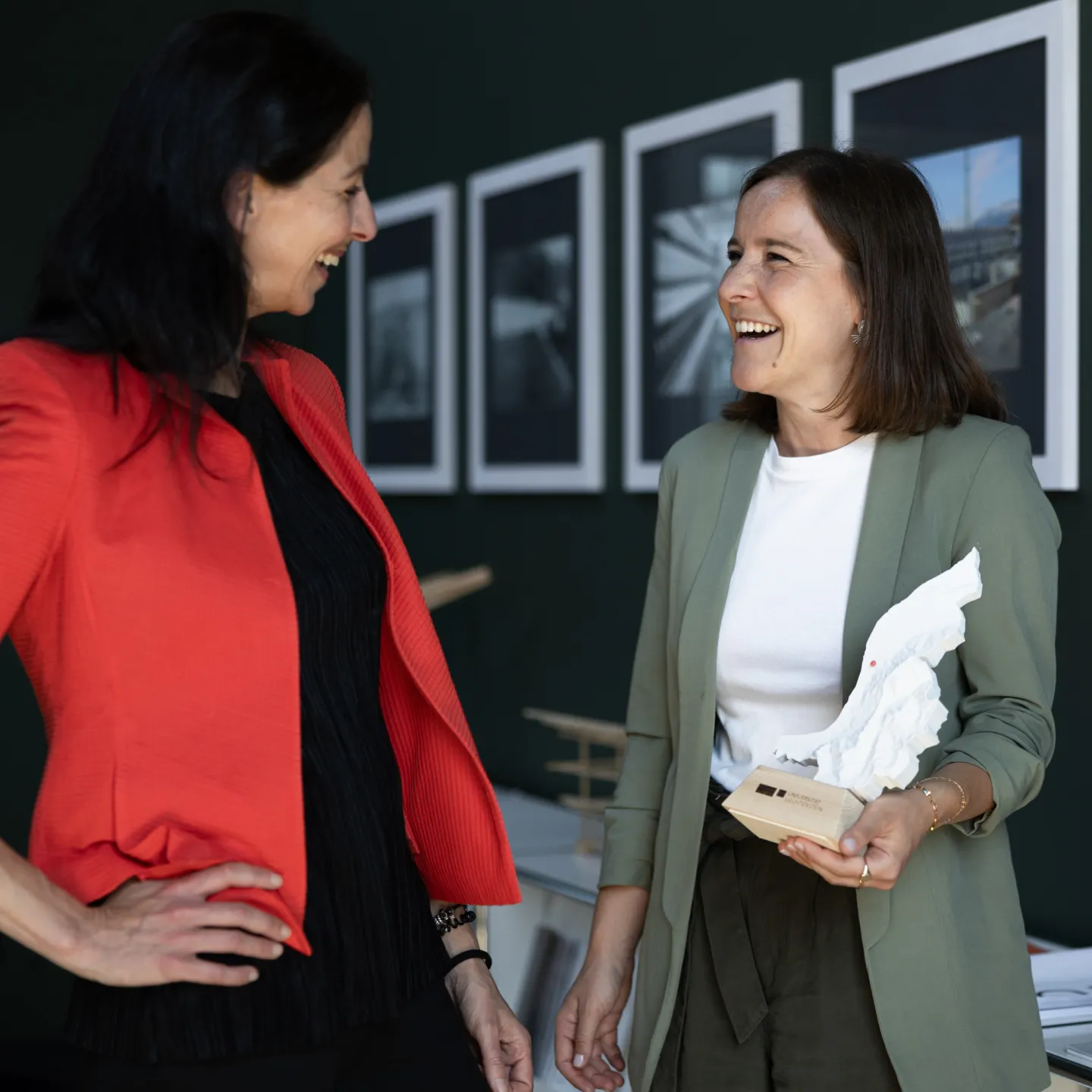 Zwei Frauen im Gespräch vor einer Bilderwand an der Universität Liechtenstein, eine hält ein Architekturmodell.