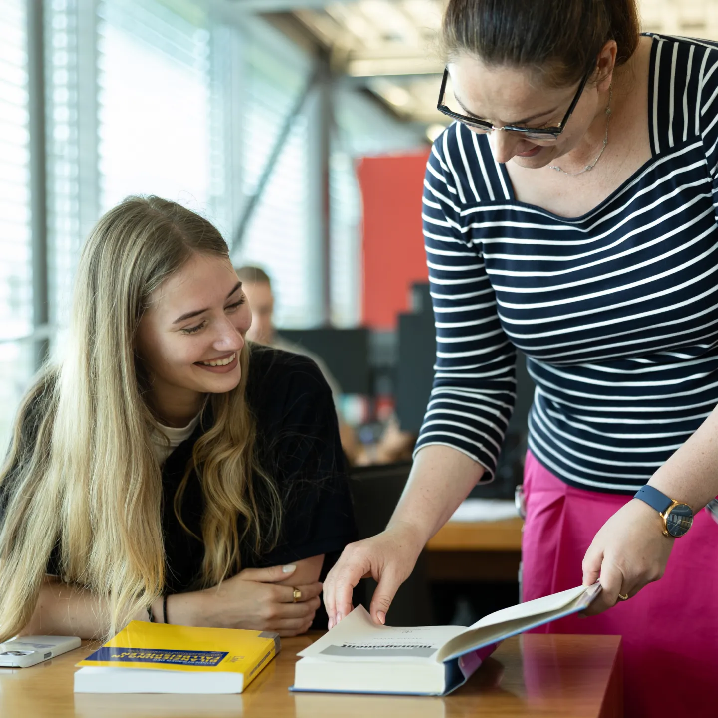 Eine Bibliotheksmitarbeiterin zeigt einer lächelnden Studentin Inhalte aus einem geöffneten Buch.