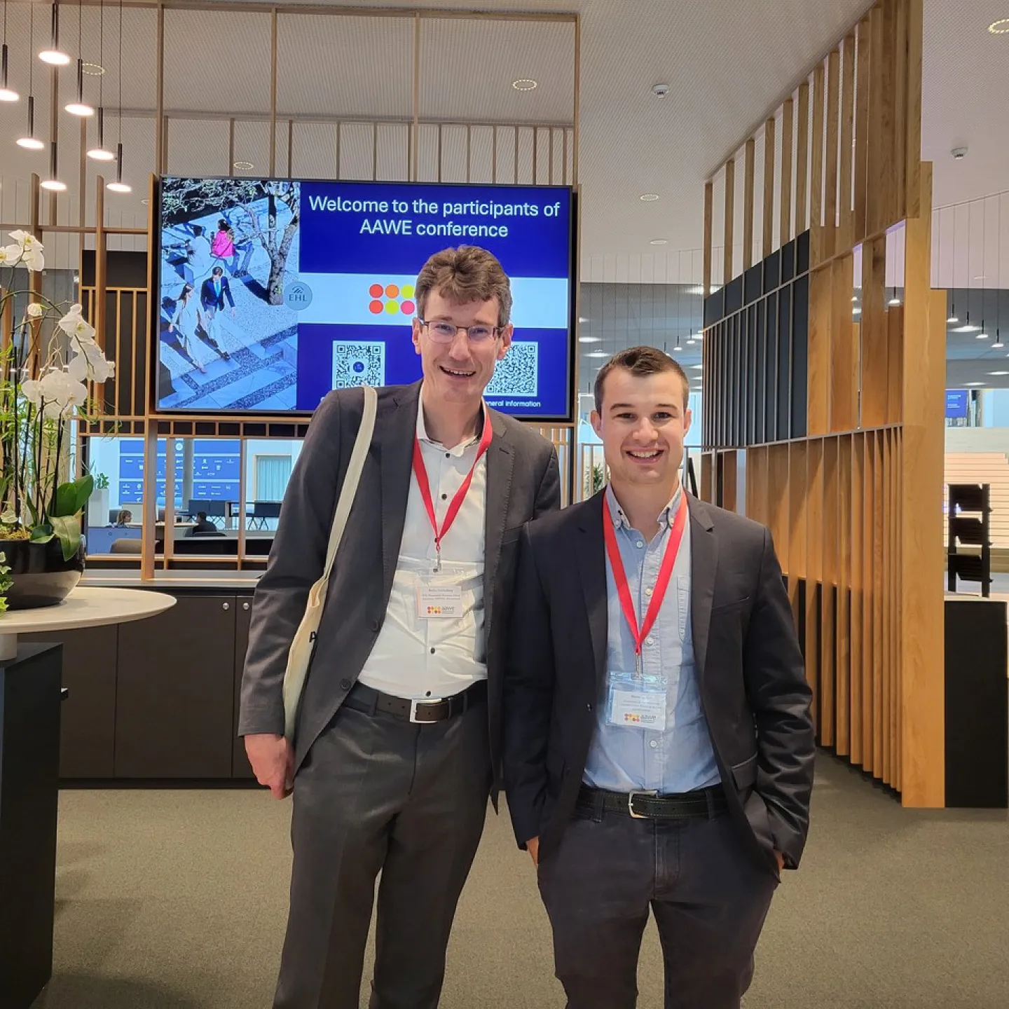 Zwei Männer in Anzügen mit roten Namensschildern stehen lächelnd in einem modernen Konferenzraum. Im Hintergrund ist ein Bildschirm mit der Aufschrift „Welcome to the participants of AAWE conference“ zu sehen. Links steht eine dekorative Pflanze auf einem Tisch.