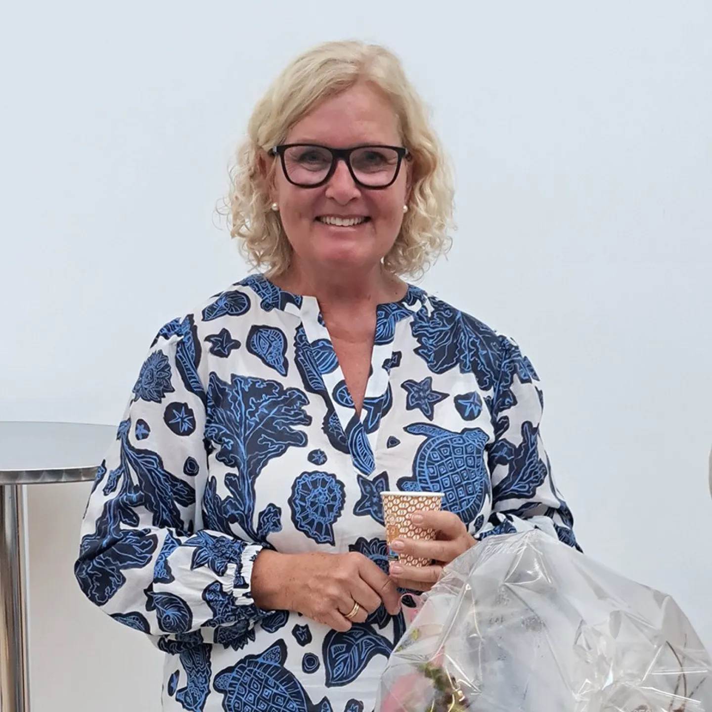 Eine lächelnde Frau mit blonden Haaren und Brille hält einen Becher in der Hand. Sie trägt ein blau-weiß gemustertes Kleid. Vor ihr steht ein Blumenstrauß in Folie verpackt.