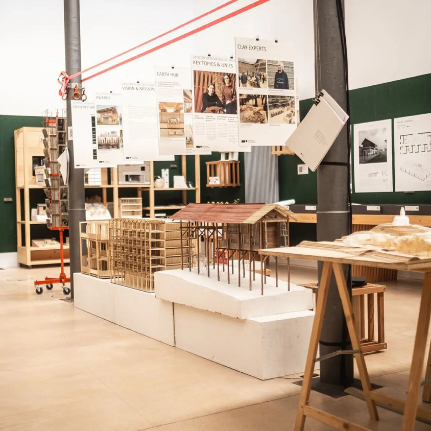 Architektur-Studienraum mit Modellen und Plakaten. Verschiedene Architekturmodelle aus Holz und Papier sind auf Tischen ausgestellt. An den Wänden hängen Plakate mit Informationen zu Projekten und Themen. Der Raum ist hell und modern gestaltet.