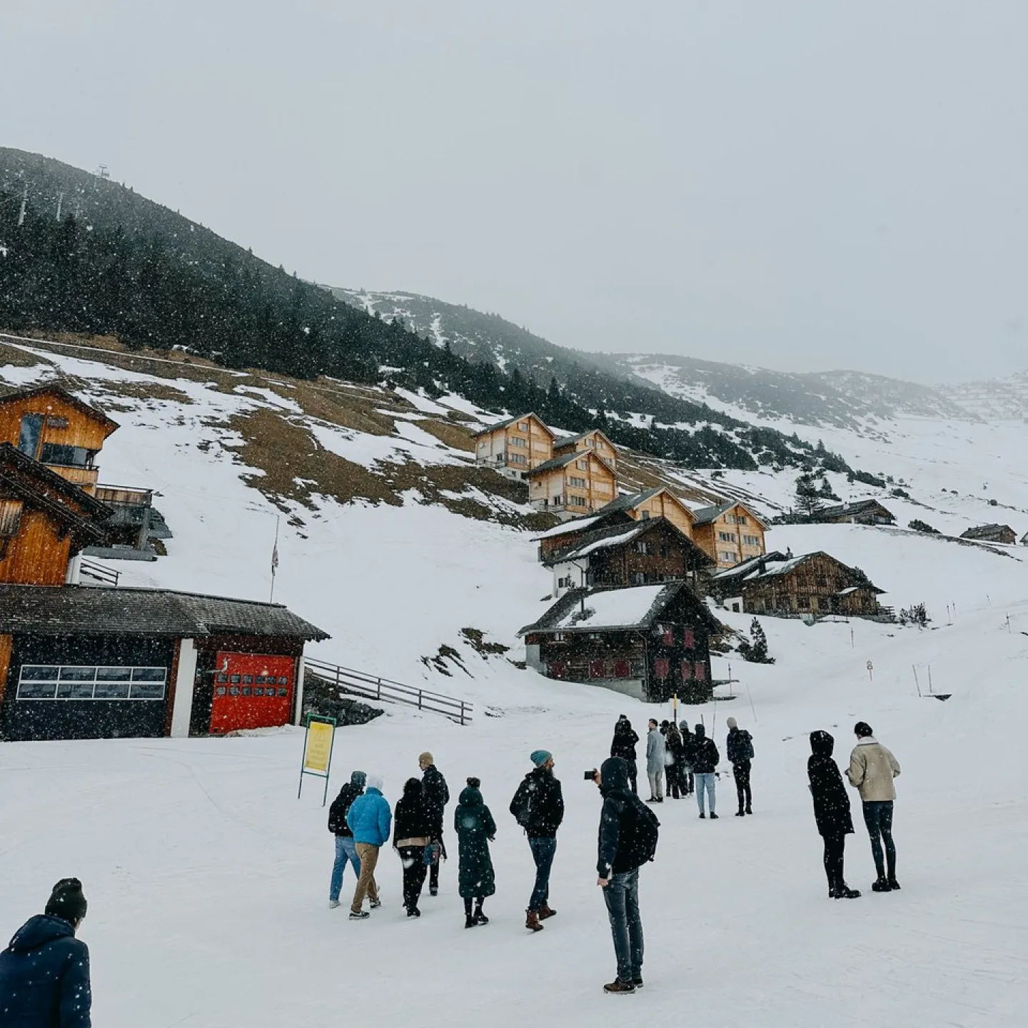 Eine Gruppe von Menschen steht in einer verschneiten Berglandschaft vor traditionellen Holzhäusern. Im Hintergrund sind schneebedeckte Hügel und Nadelbäume zu sehen. Die Szene vermittelt eine winterliche Atmosphäre und zeigt eine Verbindung von Architektur und Natur.