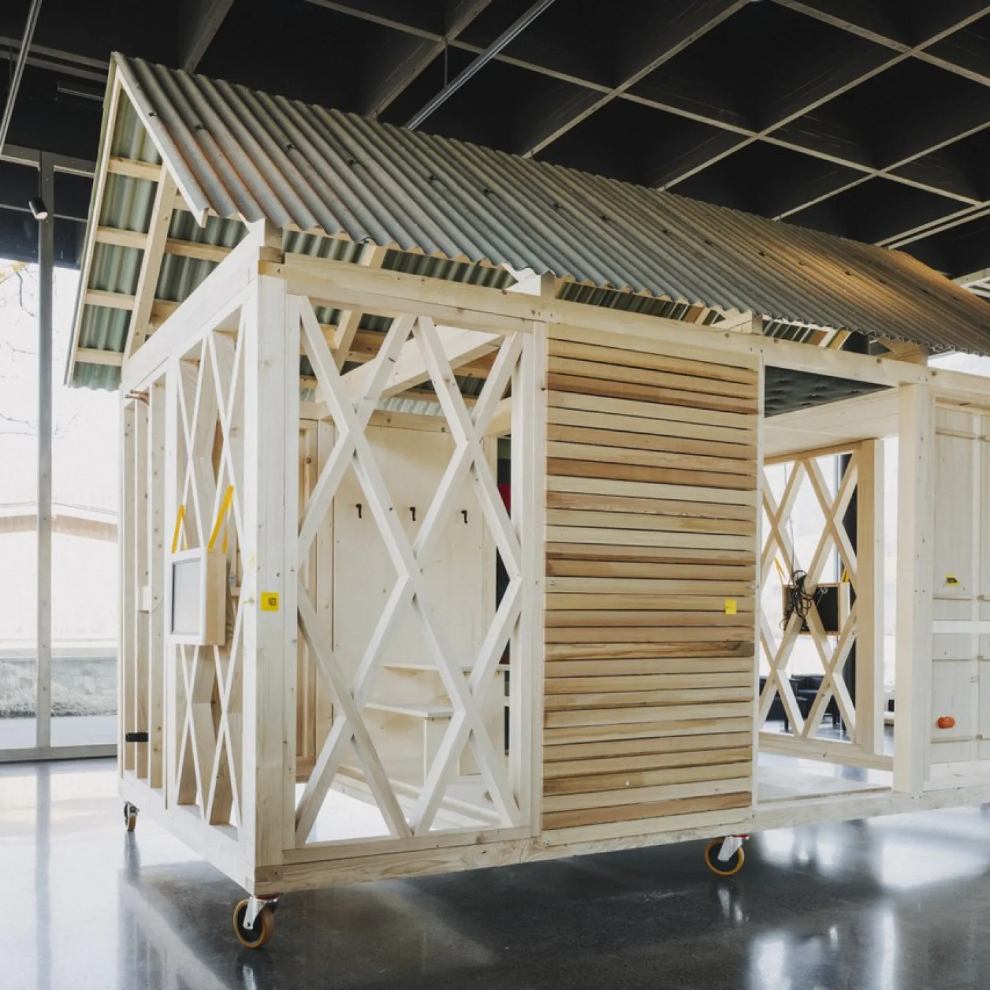 Ein einfaches, modulares Holzgebäude mit einem Wellblechdach steht auf Rollen in einem modernen, lichtdurchfluteten Raum. Das Design zeigt eine offene Struktur mit diagonalen Verstrebungen und Holzverkleidung, ideal für nachhaltiges Bauen und architektonische Bildung.