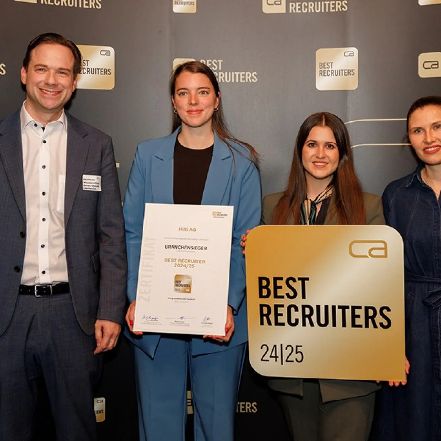 Vier Personen stehen vor einem Hintergrund mit dem Logo "Best Recruiters". Eine Person hält ein Zertifikat mit der Aufschrift "Branchen­sieger Best Recruiter 2024/25". Eine andere Person hält ein Schild mit dem gleichen Logo. Alle lächeln in die Kamera.