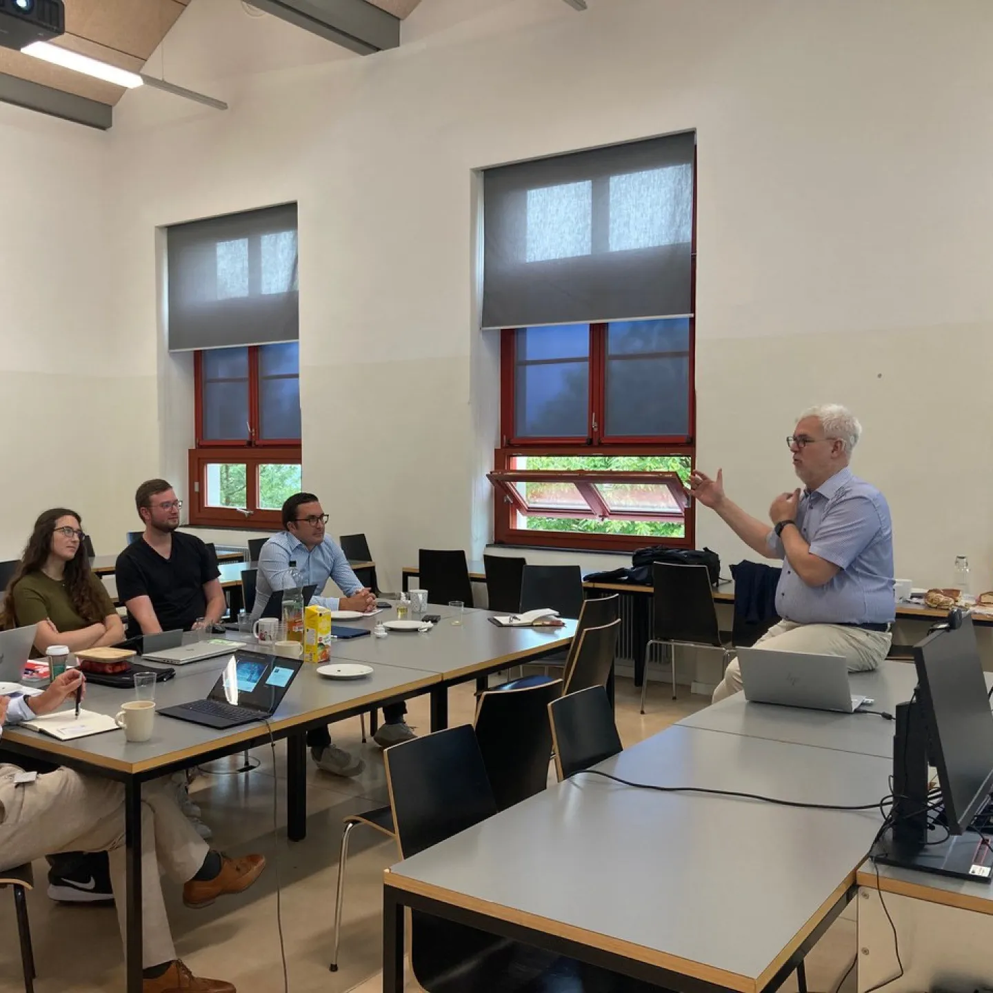 Ein Dozent hält einen Vortrag vor einer kleinen Gruppe von Personen in einem Seminarraum. Die Teilnehmenden sitzen an Tischen mit Laptops und Notizbüchern. Im Hintergrund sind Fenster und eine Leinwand zu sehen.