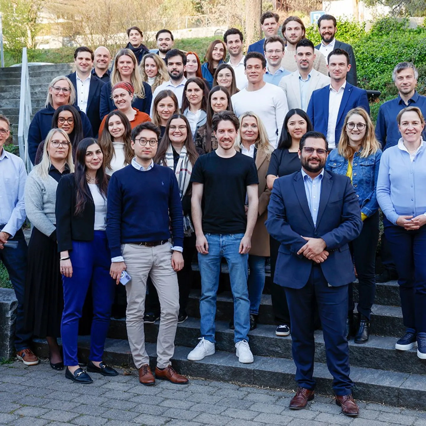 Gruppenfoto von Studierenden des Diplomstudiengangs Treuhandwesen an der Universität Liechtenstein, die auf einer Steintreppe im Freien stehen. Die Gruppe besteht aus Männern und Frauen in formeller Kleidung, umgeben von grüner Vegetation.