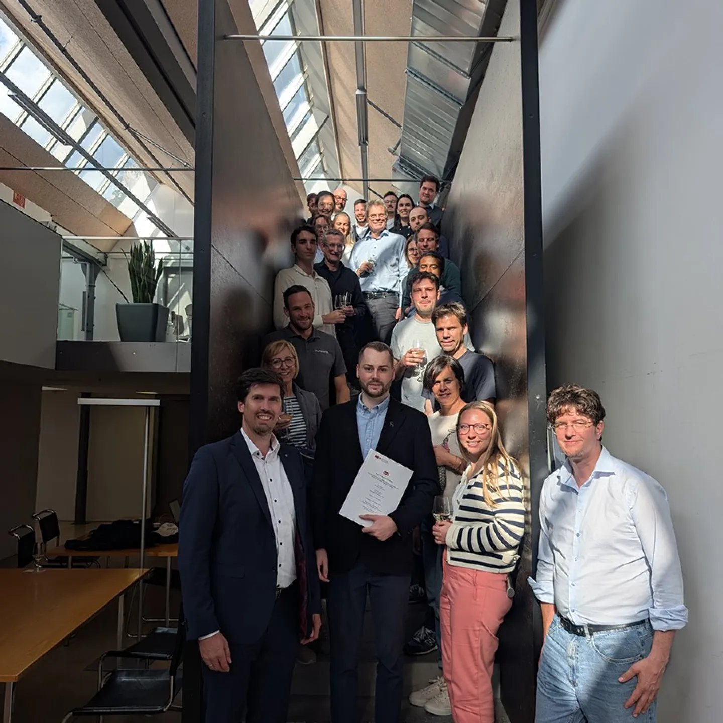 Eine Gruppe von Menschen steht auf einer Treppe in einem modernen Gebäude. Im Vordergrund hält eine Person ein Dokument in der Hand, umgeben von lächelnden Kollegen. Die Szene ist gut beleuchtet durch ein Oberlicht.