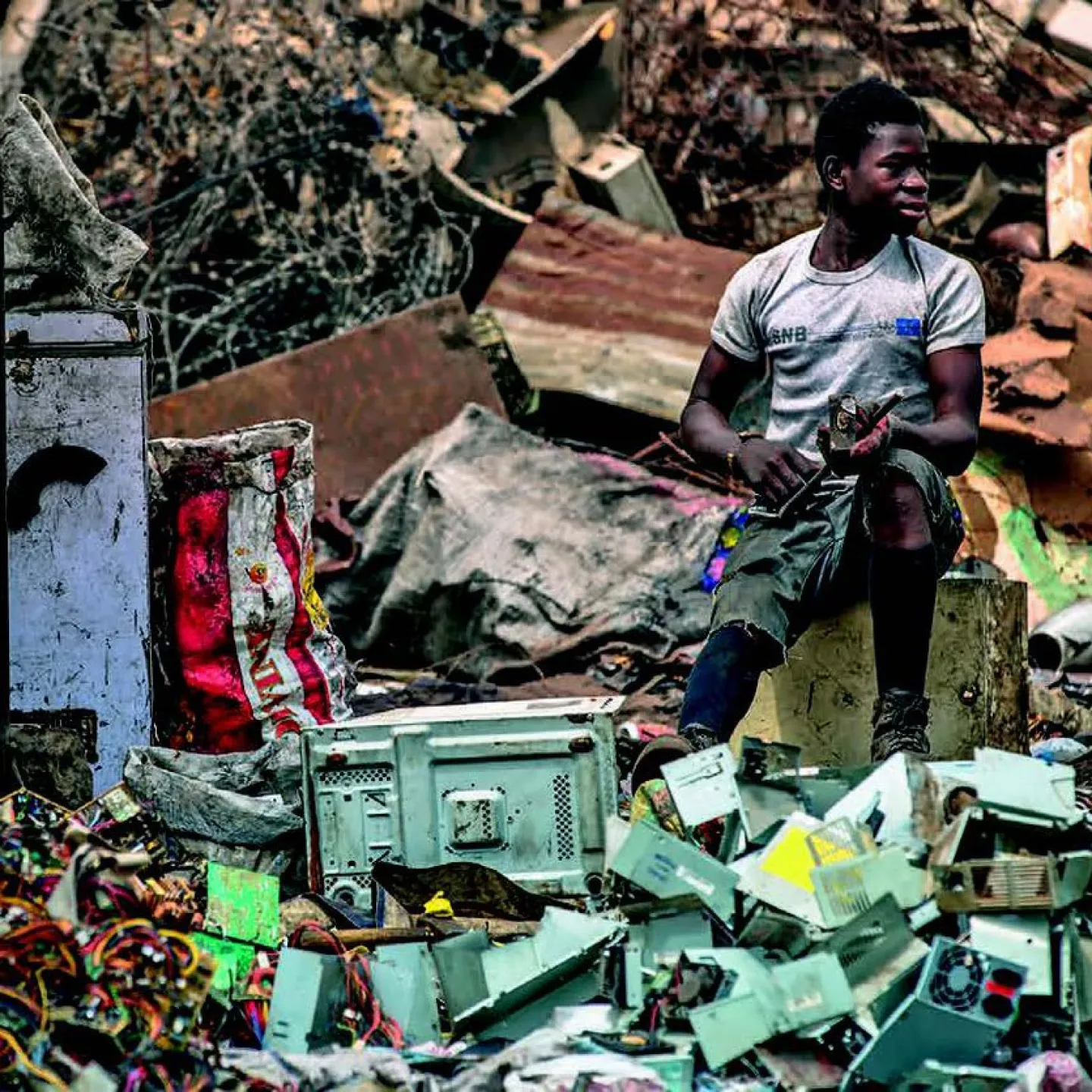 Ein Mann sitzt auf einem Hocker inmitten von Elektroschrott auf einer Müllhalde. Um ihn herum sind alte Geräte und Kabel zu sehen. Die Szene zeigt die Problematik von Elektroschrott und dessen Auswirkungen auf die Umwelt.