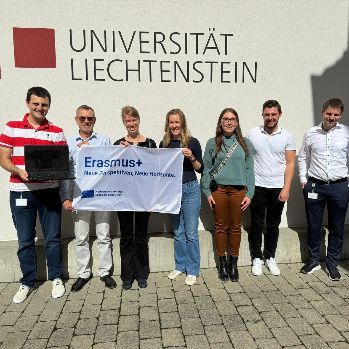 Eine Gruppe von sieben Personen steht vor einem Gebäude mit dem Schriftzug "Universität Liechtenstein". Sie halten ein Banner mit der Aufschrift "Erasmus+ Neue Perspektiven, Neue Horizonte".