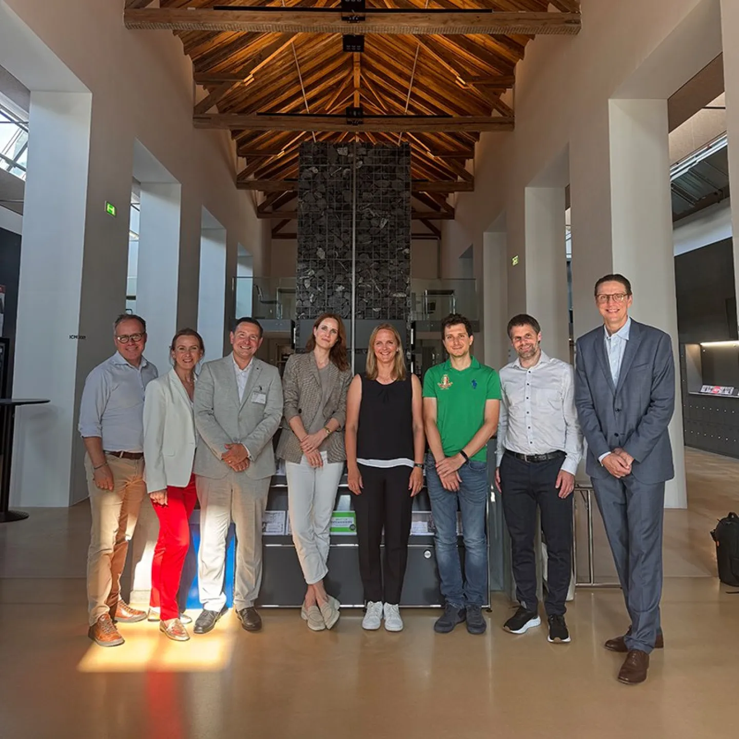 Eine Gruppe von neun Personen steht in einer modernen Halle mit Holzdach und einem Kronleuchter. Sie lächeln in die Kamera. Im Hintergrund sind hohe Fenster und eine Steinwand zu sehen.