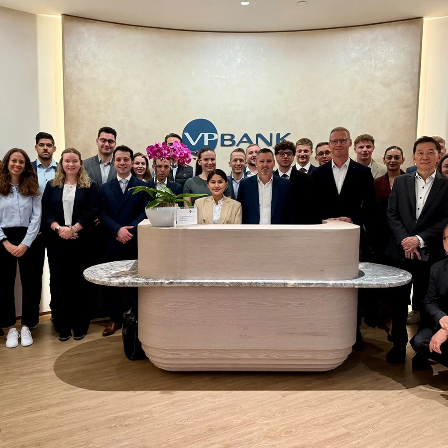 Gruppe von Studierenden und Fachleuten in formeller Kleidung steht in der Lobby der VP Bank. Im Hintergrund ist das Logo der Bank sichtbar. Die Szene vermittelt eine professionelle und lernorientierte Atmosphäre.