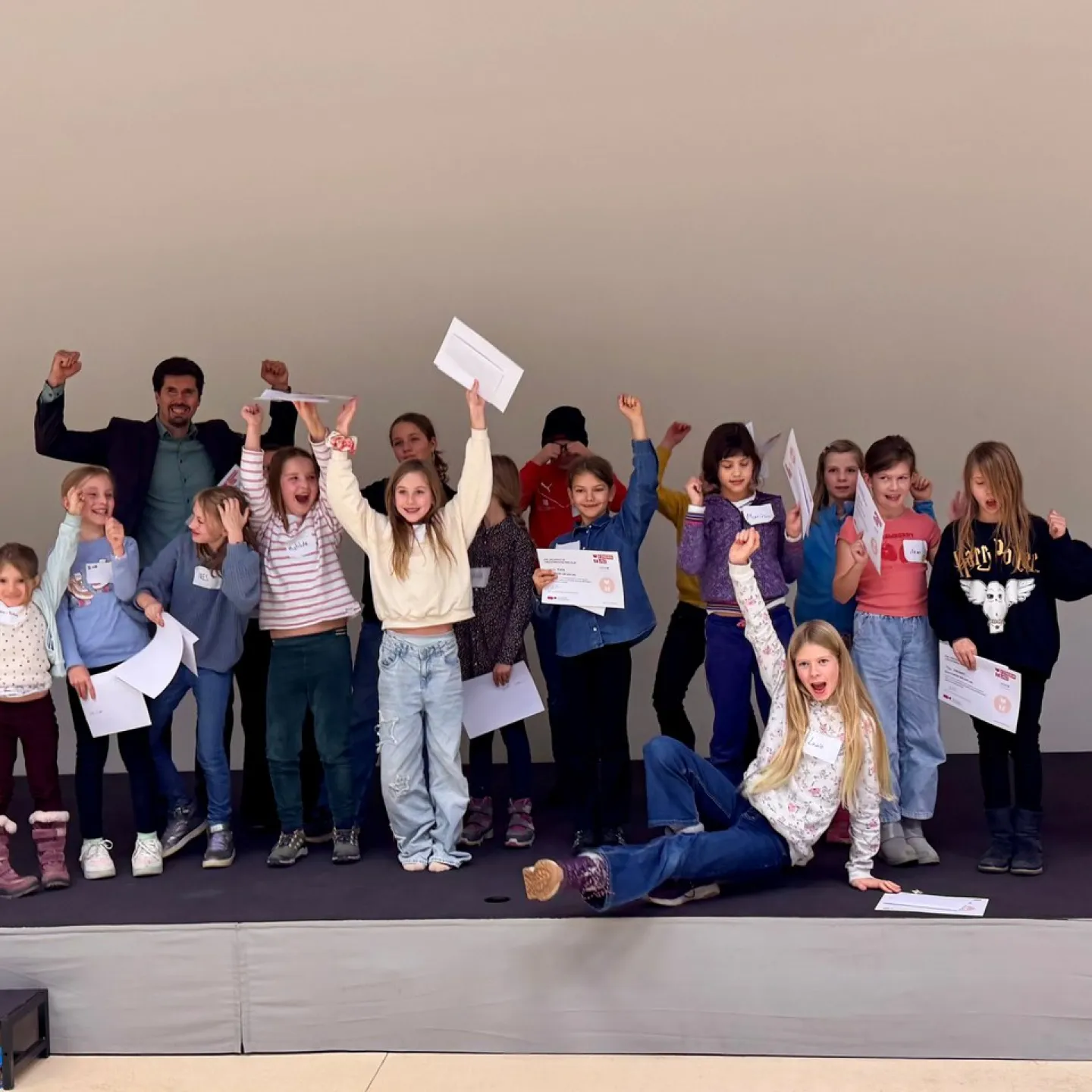 Eine Gruppe von Kindern und einem Erwachsenen steht auf einer Bühne und hält stolz Zertifikate hoch. Die Kinder wirken fröhlich und begeistert. Sie nehmen an einem Fundraising-Workshop an der Universität Liechtenstein teil.