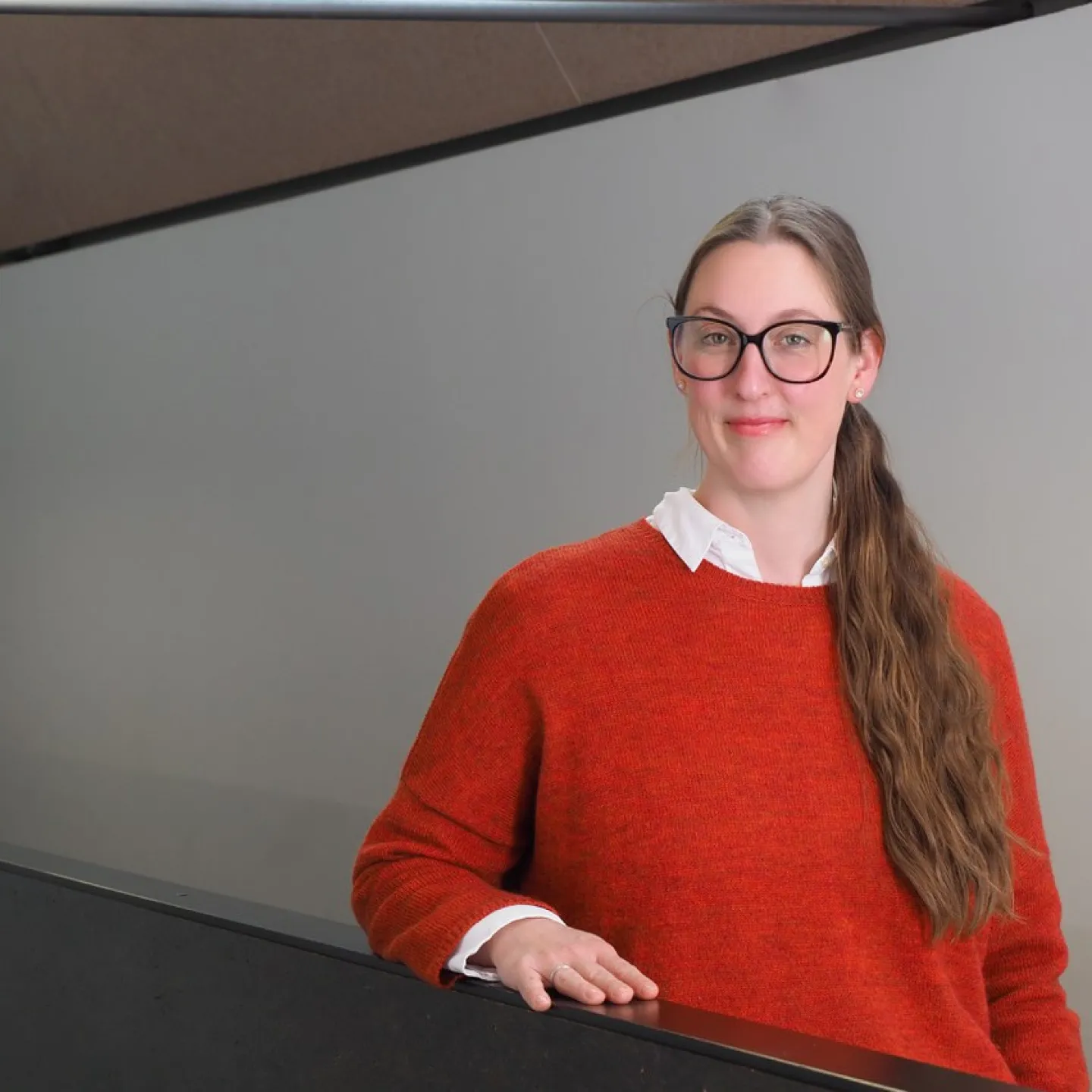 Eine Person mit Brille und langem Haar steht in einem modernen Gebäude und trägt einen roten Pullover. Sie lächelt leicht und lehnt sich an ein Geländer.