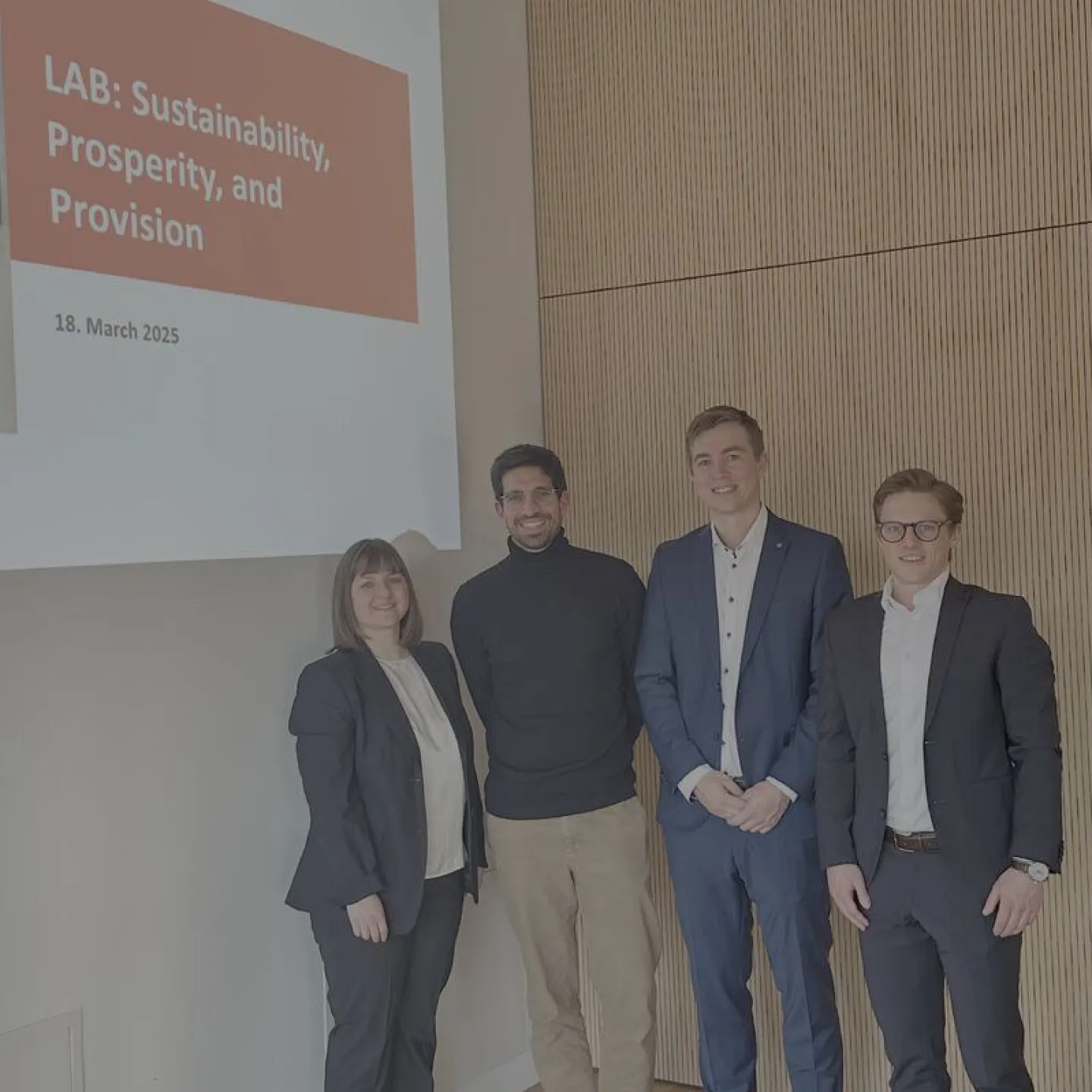 Vier Personen stehen lächelnd vor einer Präsentationsfolie mit der Aufschrift "LAB: Sustainability, Prosperity, and Provision, 18. March 2025". Sie befinden sich in einem modernen Raum mit Holzvertäfelung.