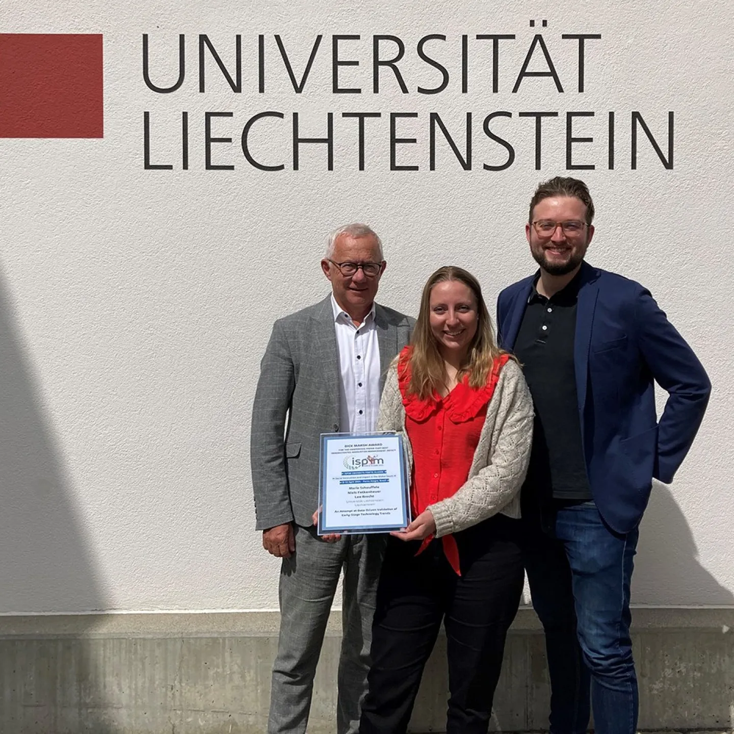 Drei Personen stehen vor einer Wand mit dem Logo der Universität Liechtenstein. Eine Person hält eine Auszeichnung in den Händen.