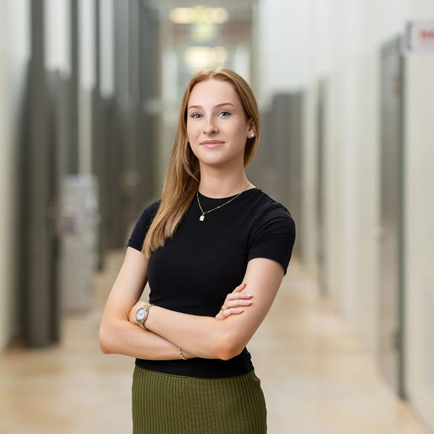 Eine junge Frau steht selbstbewusst mit verschränkten Armen in einem modernen Flur. Sie trägt ein schwarzes Oberteil und einen grünen Rock.