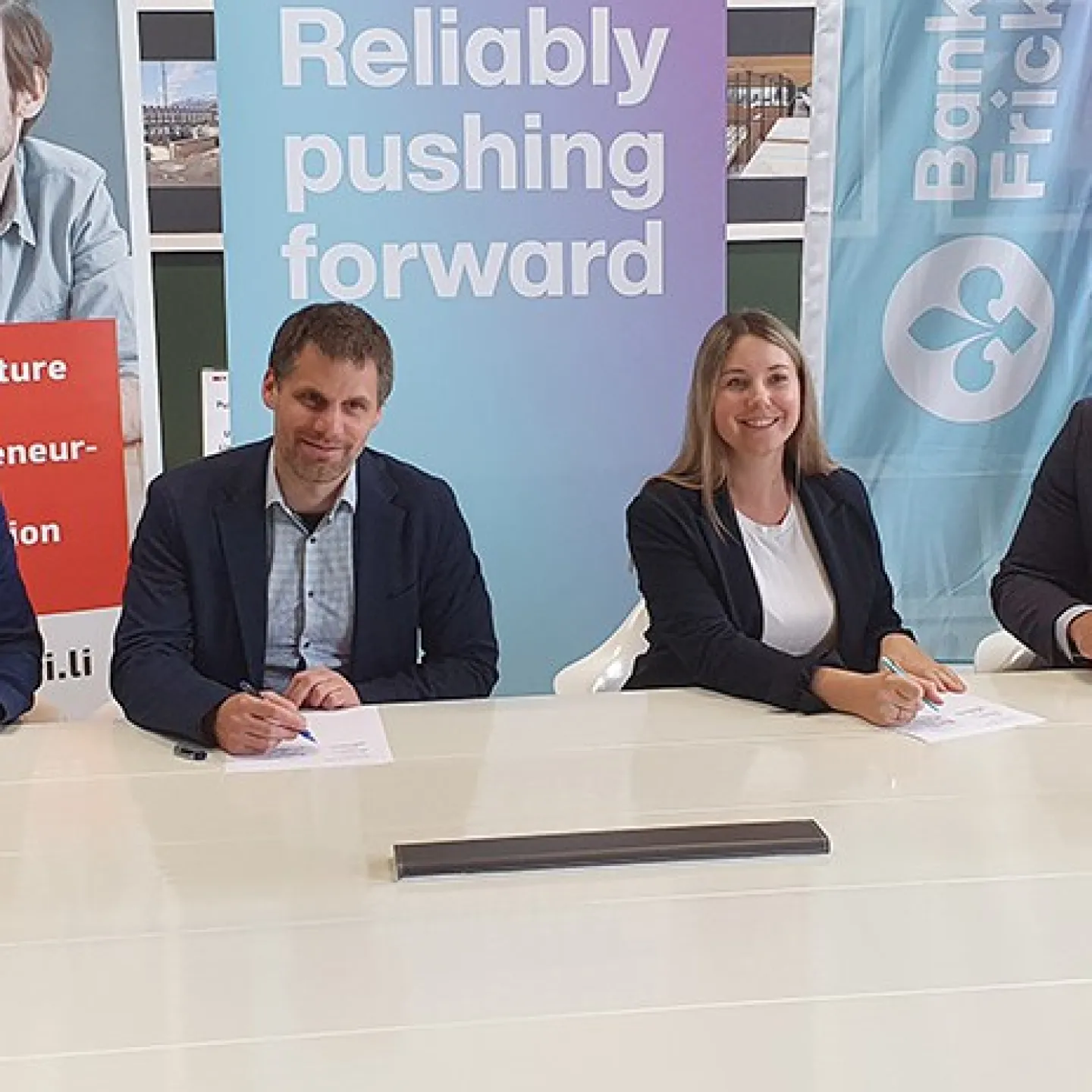 Vier Personen sitzen an einem Konferenztisch und unterzeichnen Dokumente. Im Hintergrund sind Banner der Universität Liechtenstein und Bank Frick zu sehen. Die Szene symbolisiert die Verlängerung der Kooperation zwischen der Universität und der Bank.
