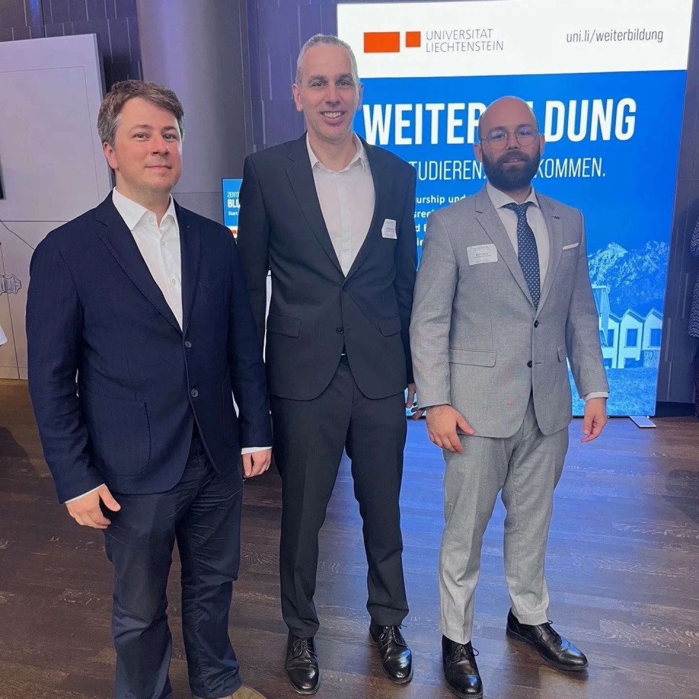 Drei Männer in Anzügen stehen vor einem Schild der Universität Liechtenstein. Im Hintergrund ist ein Banner mit der Aufschrift "Weiterbildung" zu sehen.