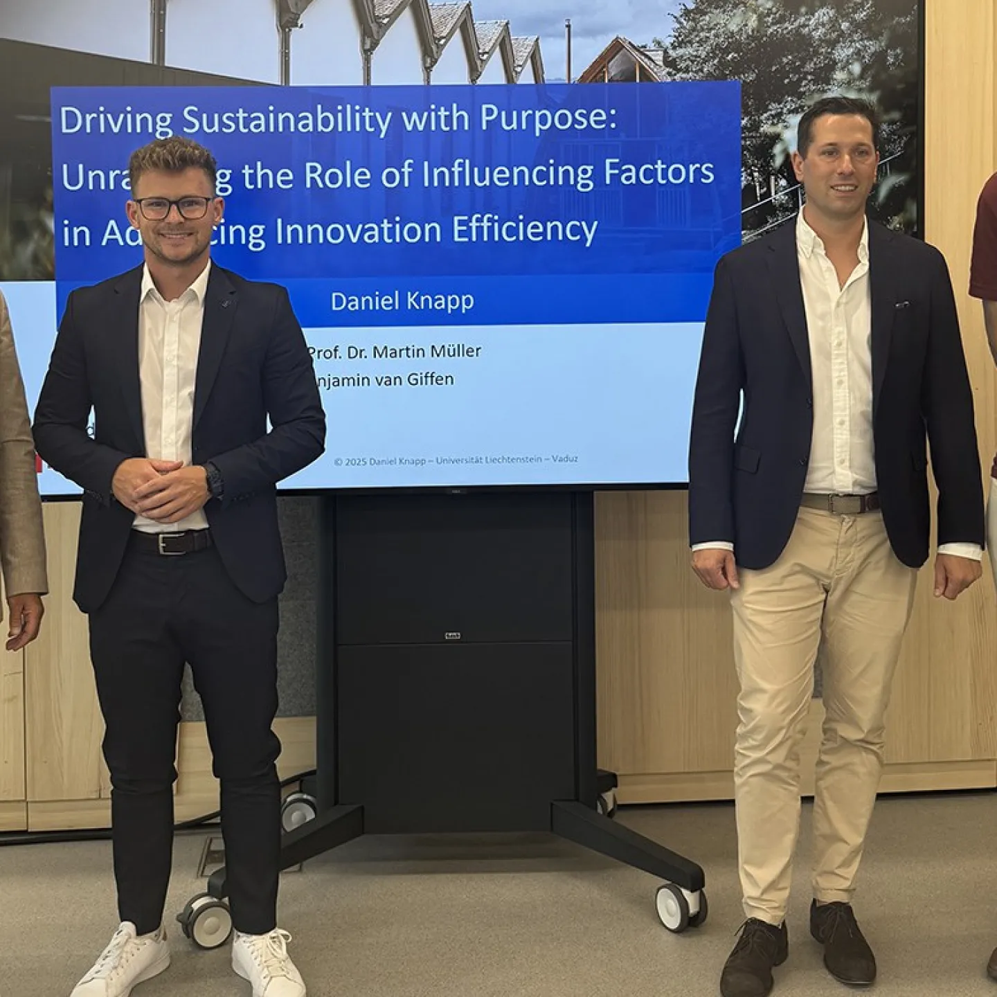 Vier Männer stehen vor einem Bildschirm, der eine Präsentation mit dem Titel „Driving Sustainability with Purpose“ zeigt. Sie tragen Business-Kleidung und lächeln in die Kamera. Der Hintergrund ist ein moderner Innenraum.