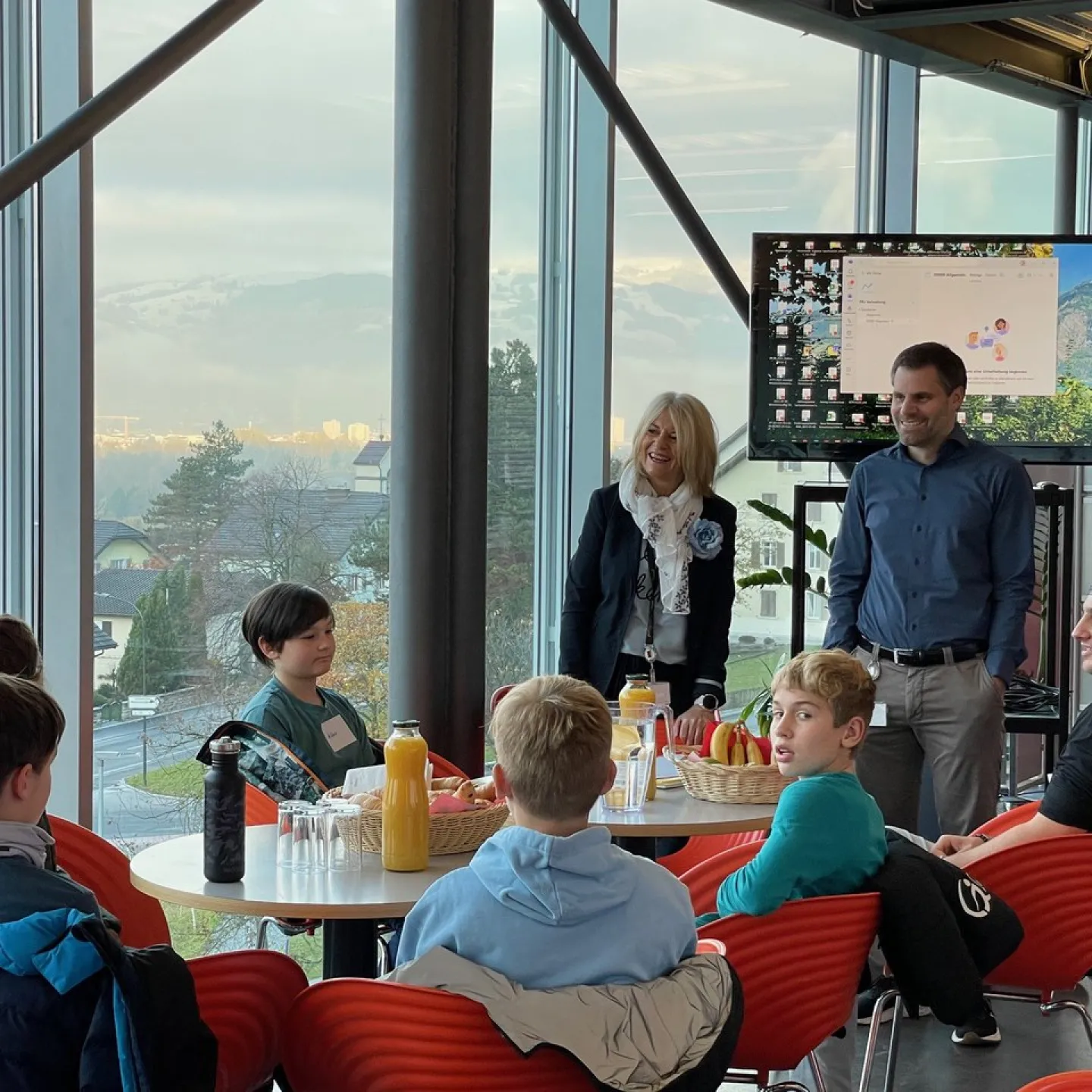 Eine Gruppe von Kindern sitzt an einem Tisch in einem modernen Raum mit großen Fenstern. Zwei Erwachsene stehen lächelnd daneben. Im Hintergrund ist ein Bildschirm zu sehen. Die Szene wirkt freundlich und einladend.