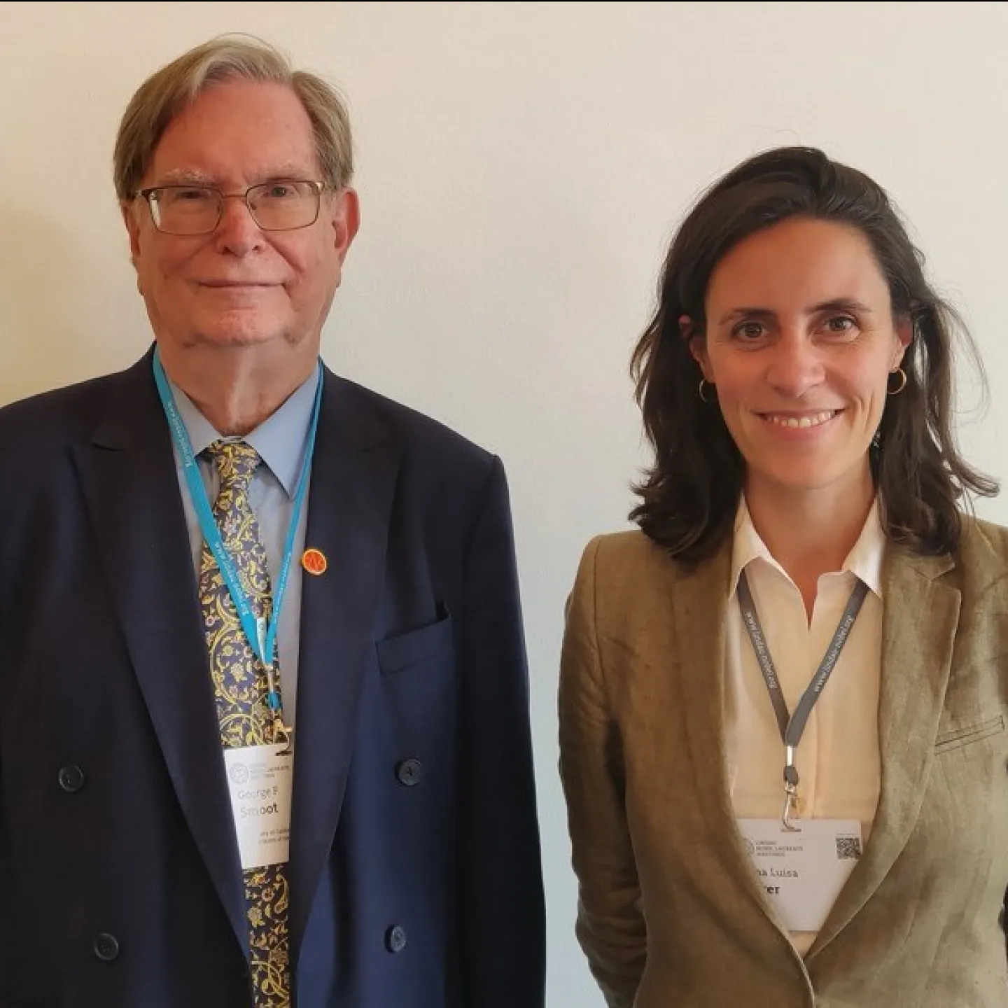 Zwei Personen stehen nebeneinander und lächeln in die Kamera. Beide tragen Namensschilder und sind formell gekleidet.