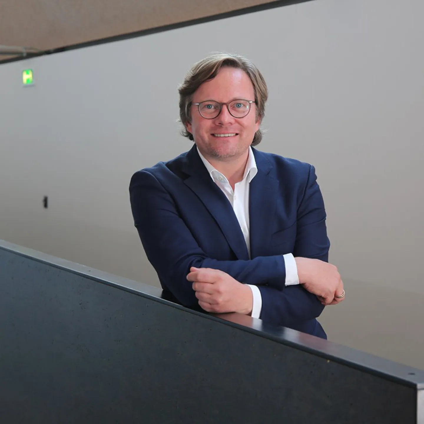 Ein Mann in einem blauen Anzug lehnt lächelnd an einem Geländer in einem modernen Gebäude.