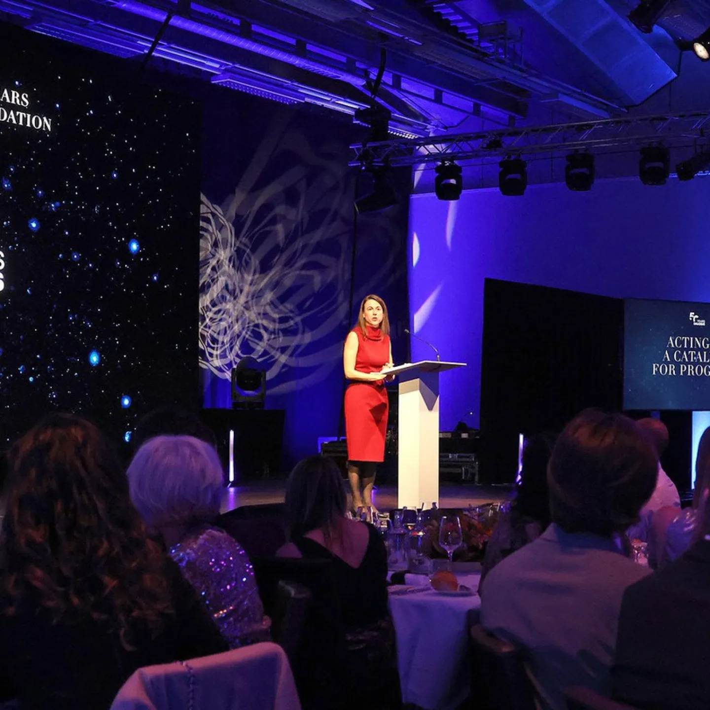 Eine Rednerin in rotem Kleid steht an einem Podium auf einer Bühne, umgeben von Publikum. Im Hintergrund ist eine Leinwand mit der Aufschrift "Celebrating 50 Years of the Onassis Foundation" zu sehen. Die Veranstaltung findet in einem festlich beleuchteten Raum statt.