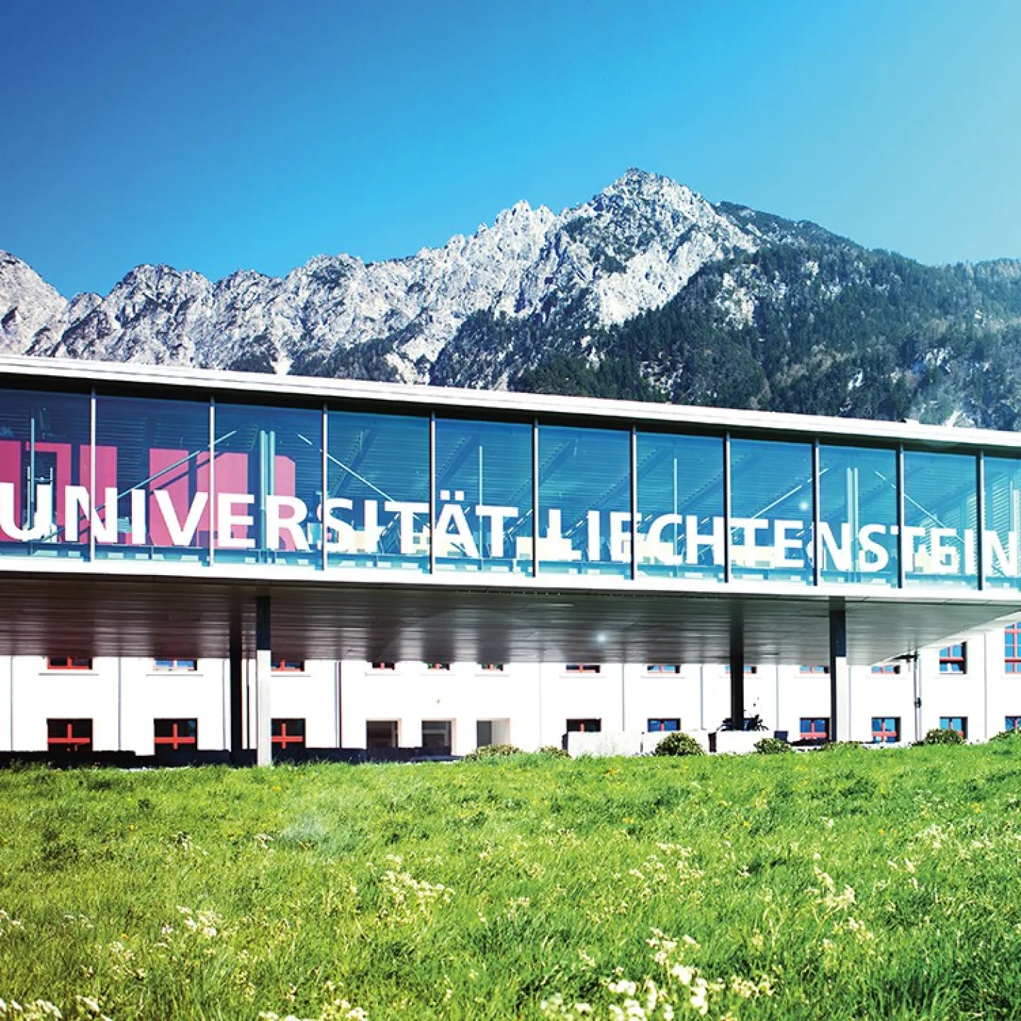 Außenansicht der Universität Liechtenstein mit modernem Glasgebäude und Bergkulisse im Hintergrund, umgeben von grüner Wiese.