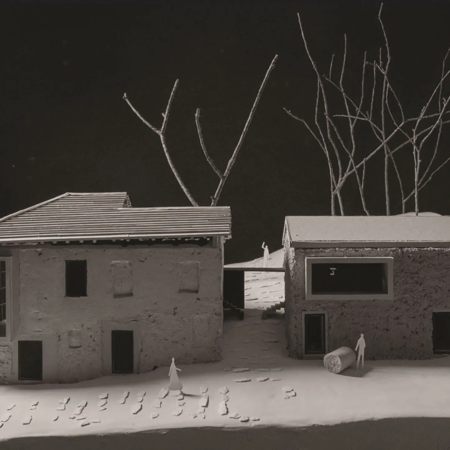 Modell eines architektonischen Projekts mit zwei Gebäuden, umgeben von kahlen Bäumen. Das linke Gebäude hat ein geneigtes Dach, während das rechte ein Flachdach aufweist. Kleine Figuren und Fußspuren sind auf dem Boden sichtbar, was eine menschliche Präsenz andeutet.