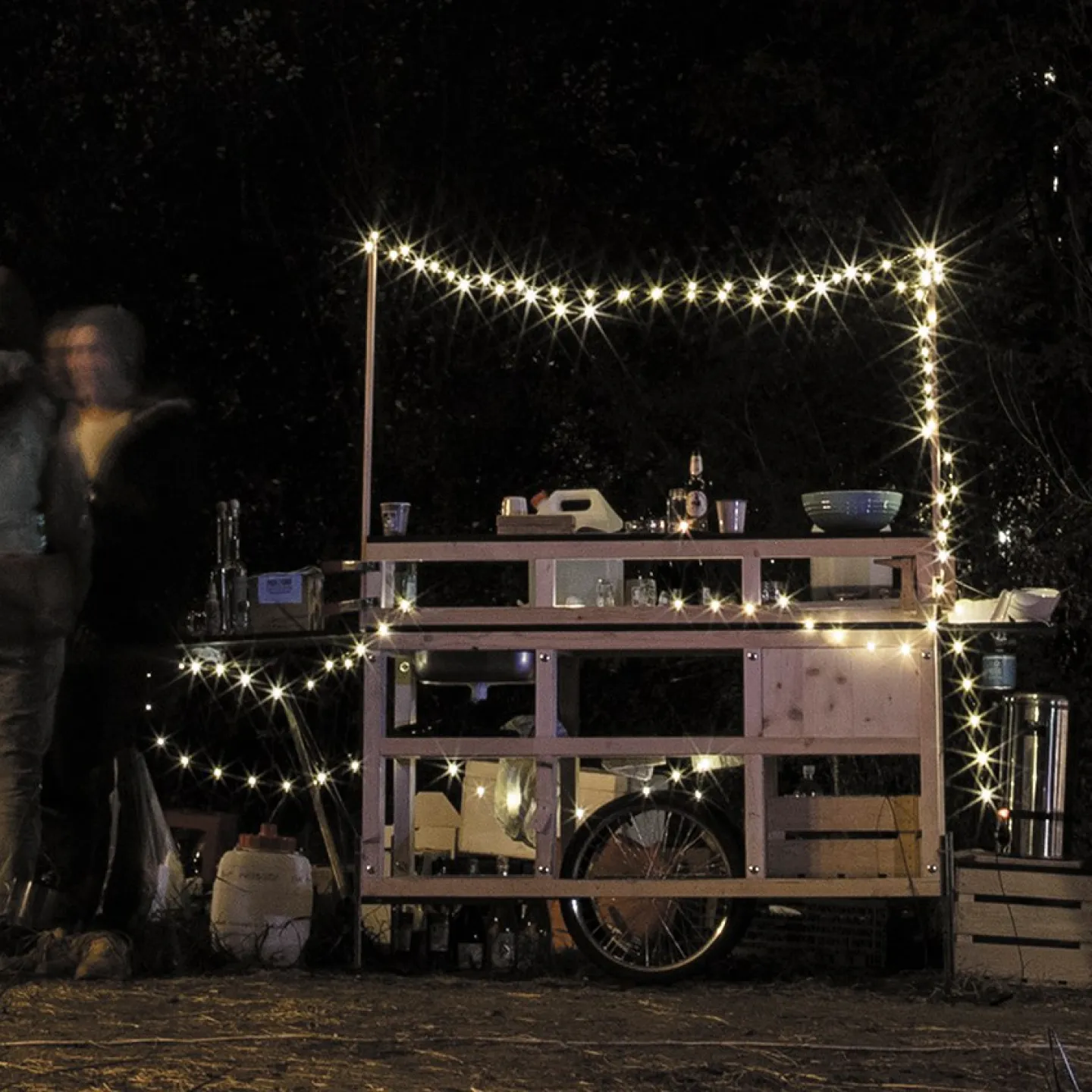 Eine mobile Holzbar, dekoriert mit Lichterketten, steht im Freien bei Nacht. Mehrere Personen stehen links neben der Bar und unterhalten sich. Die Szene vermittelt eine gemütliche und einladende Atmosphäre.