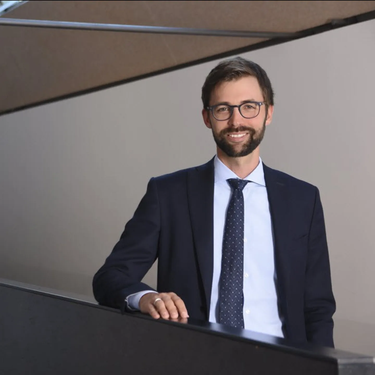 Ein Mann in Anzug und Krawatte steht lächelnd in einem modernen Gebäude mit großen Fenstern.