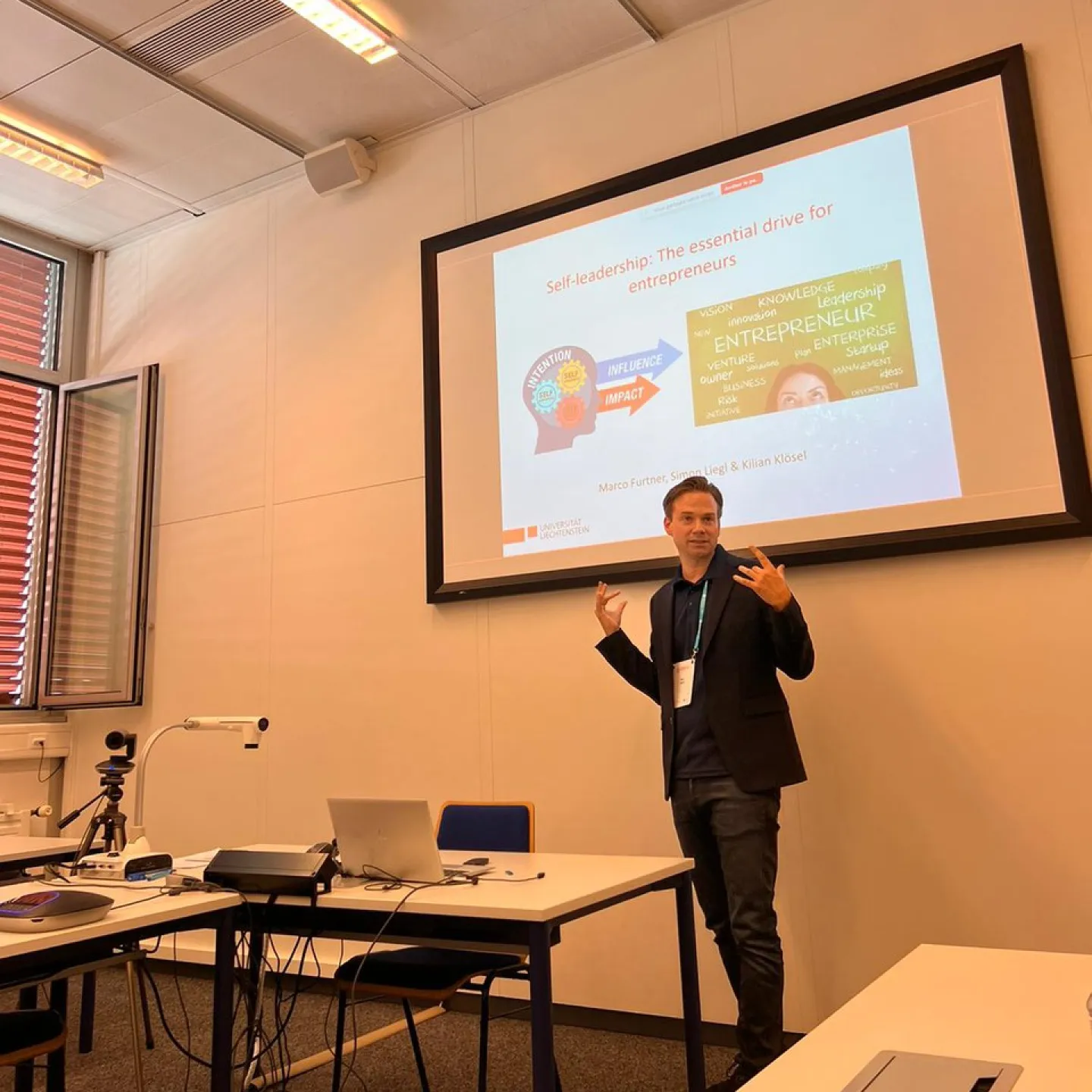 Ein Vortragender präsentiert in einem Seminarraum ein Paper über Selbstführung für Unternehmer. Im Hintergrund ist eine Leinwand mit einer Präsentationsfolie zu sehen. Tische mit Laptops und Kamera sind im Vordergrund.