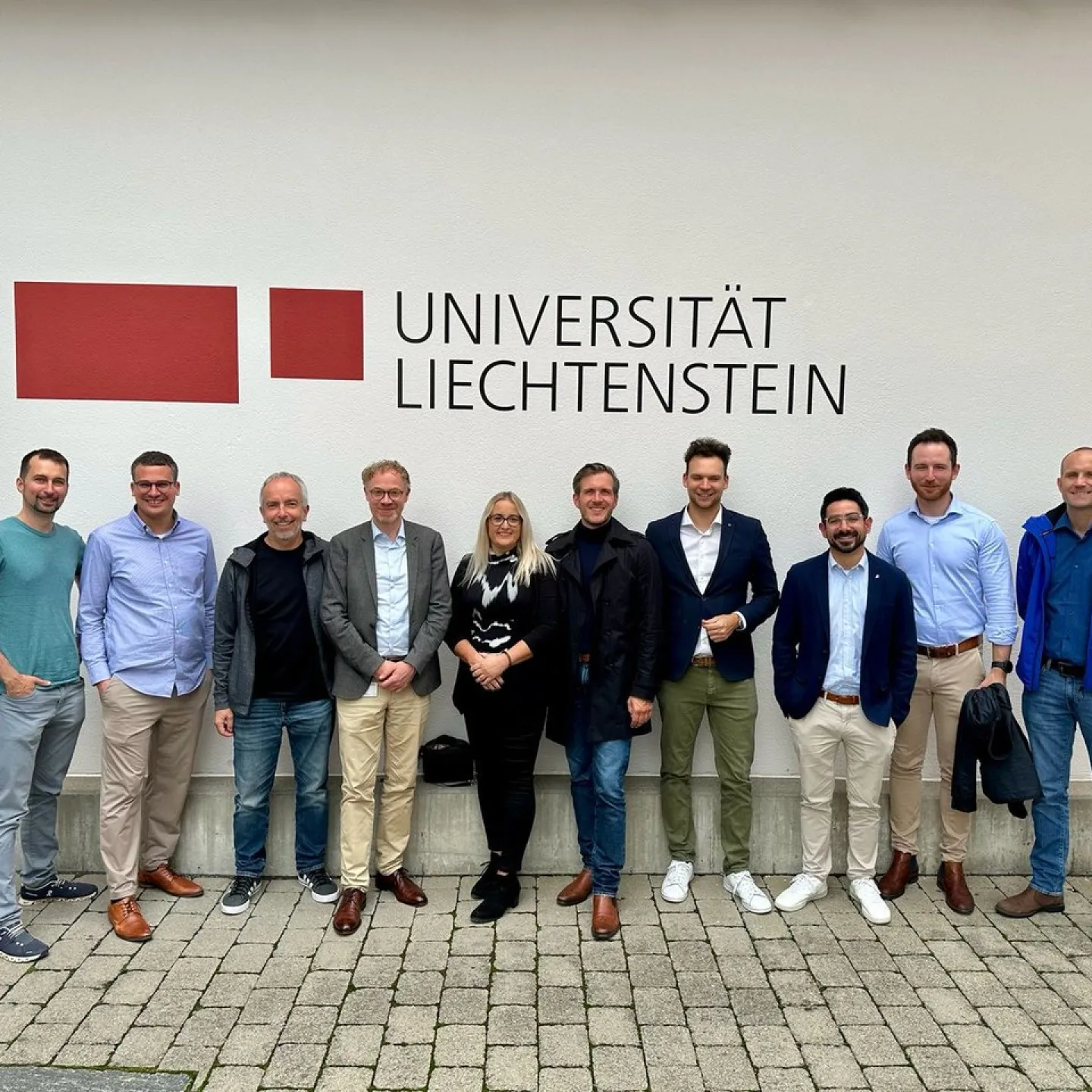 Gruppe von zehn Personen steht lächelnd vor einer Wand mit dem Logo der Universität Liechtenstein. Sie tragen unterschiedliche Freizeit- und Businesskleidung.