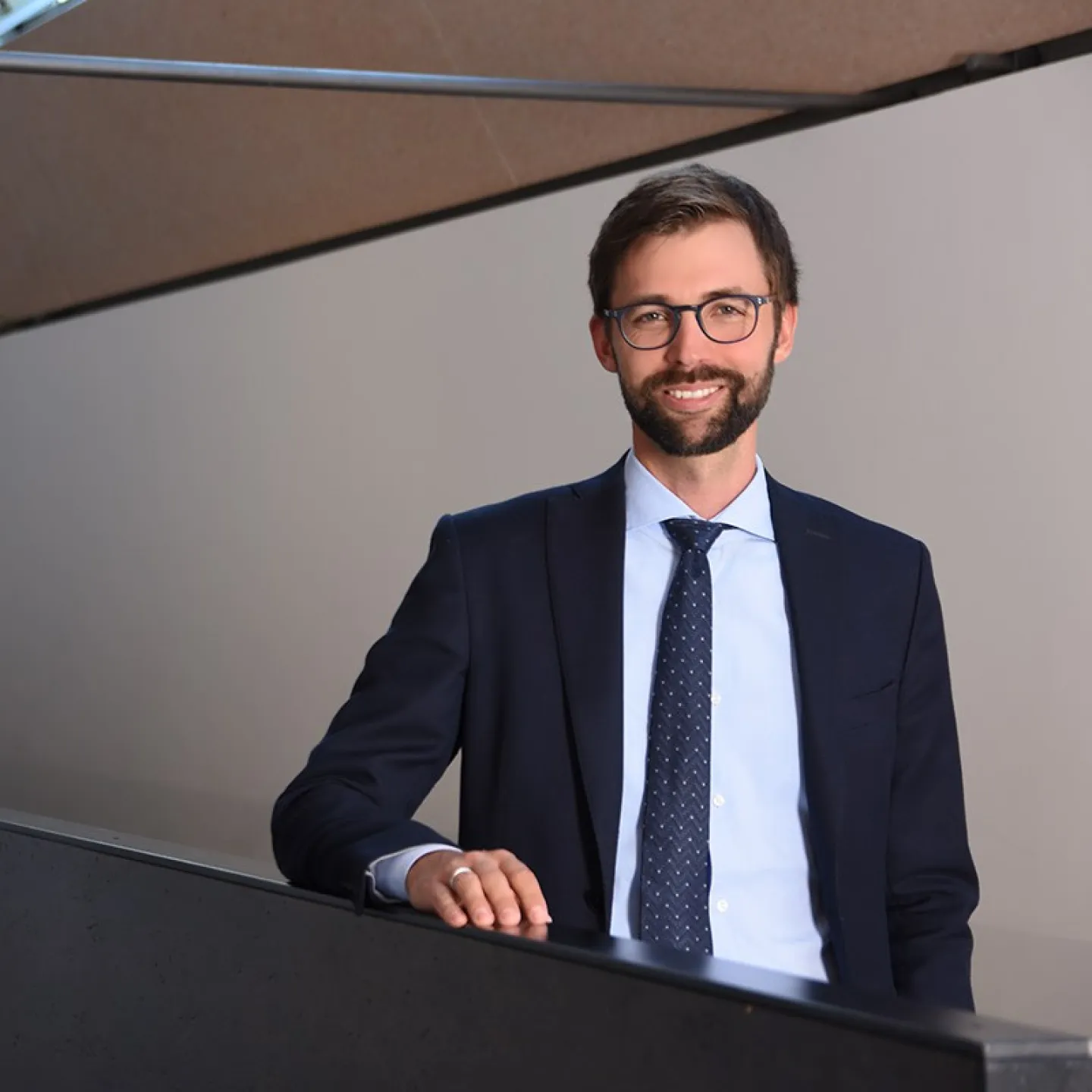 Ein Mann in Anzug und Krawatte steht lächelnd in einem modernen Gebäude mit schrägen Decken und großen Fenstern.