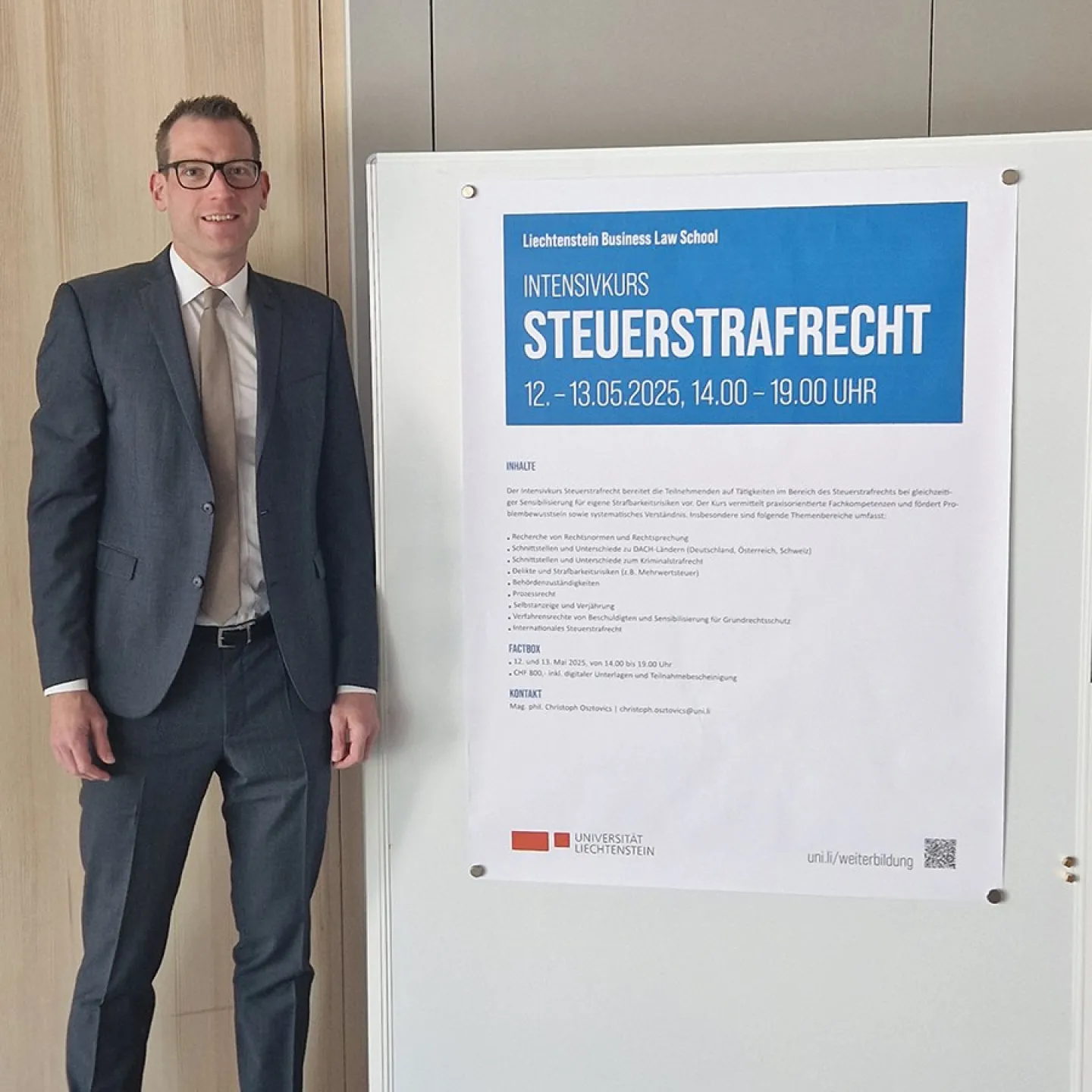 Ein Mann in Anzug steht neben einem Plakat, das den Intensivkurs „Steuerstrafrecht“ der Universität Liechtenstein ankündigt. Das Plakat enthält Informationen zu Datum, Uhrzeit und Inhalten des Kurses.