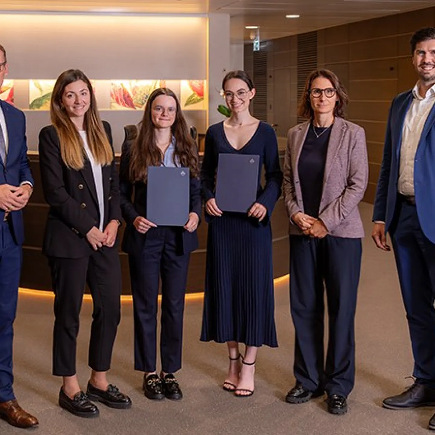 Eine Gruppe von sechs Personen steht in einem modernen Bürogebäude. Zwei Frauen in der Mitte halten Zertifikate und lächeln. Sie werden von vier weiteren Personen flankiert, die ebenfalls lächeln. Die Szene vermittelt eine feierliche Atmosphäre, möglicherweise im Rahmen einer Stipendienverleihung an der Universität Liechtenstein.
