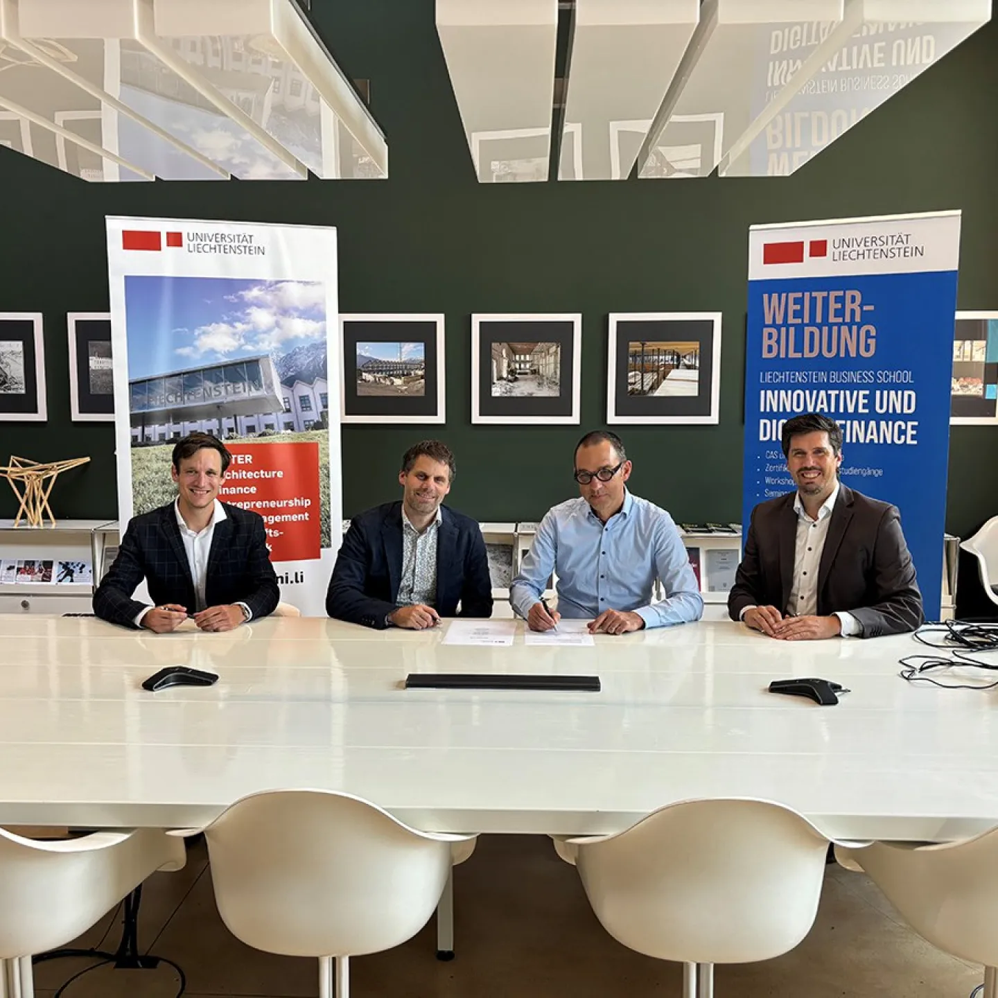 Vier Männer sitzen an einem Tisch in einem modernen Konferenzraum der Universität Liechtenstein. Im Hintergrund sind Banner mit dem Logo der Universität und Informationen zur Weiterbildung sichtbar.