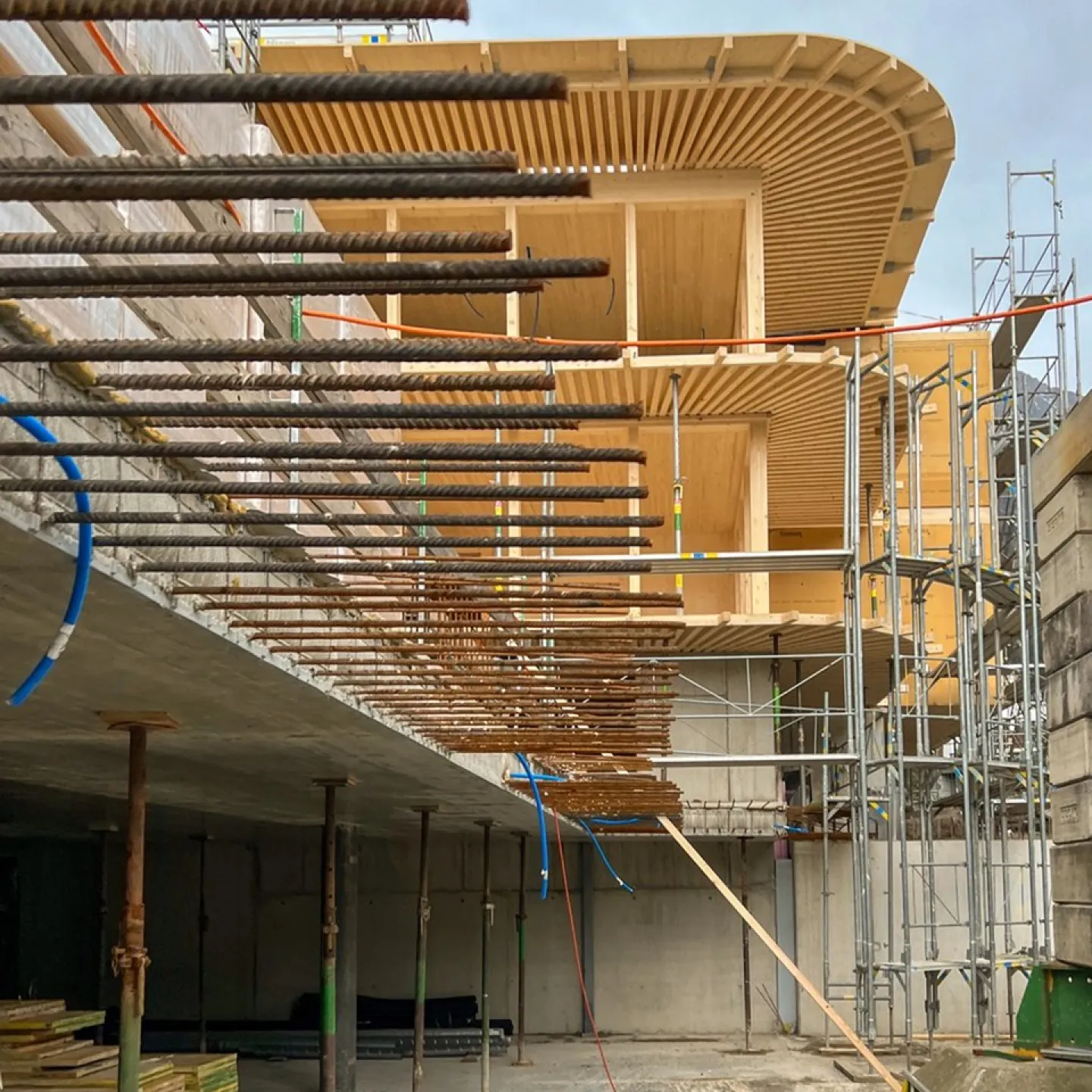 Baustelle eines modernen Gebäudes mit sichtbaren Stahlträgern und Holzverkleidung, umgeben von Gerüsten, unter bewölktem Himmel.