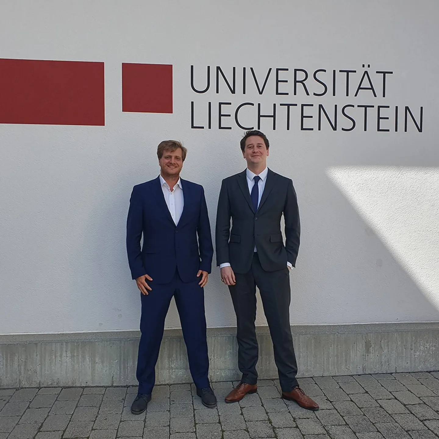 Zwei Personen in Anzügen stehen vor einer Wand mit dem Logo der Universität Liechtenstein.