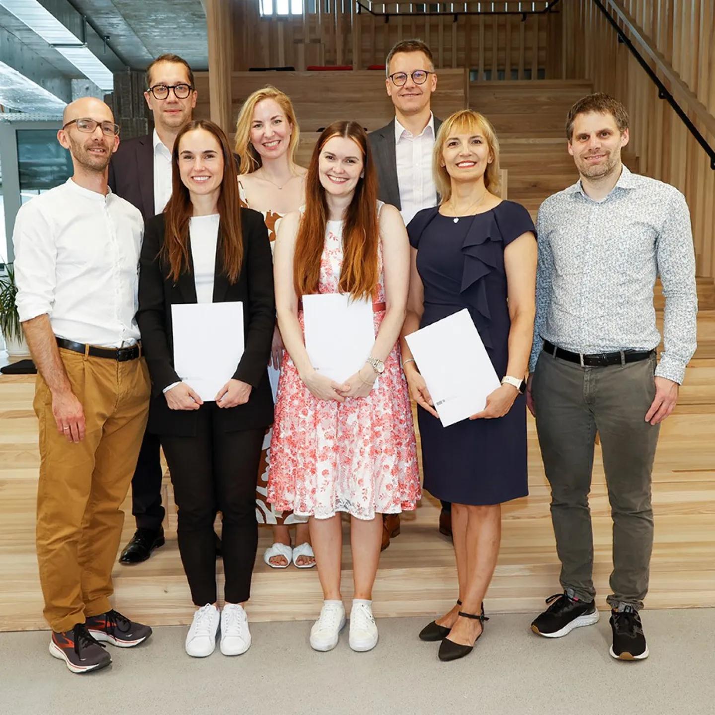 Eine Gruppe von acht Personen steht lächelnd in einem modernen Innenraum. Drei Personen in der vorderen Reihe halten Zertifikate in den Händen. Die Umgebung wirkt hell und einladend mit Holzelementen und Pflanzen.