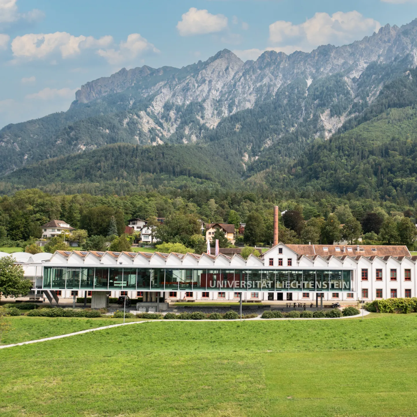 Frontansicht der Universität Liechtenstein mit markanter Glasfassade und grüner Wiese im Vordergrund.