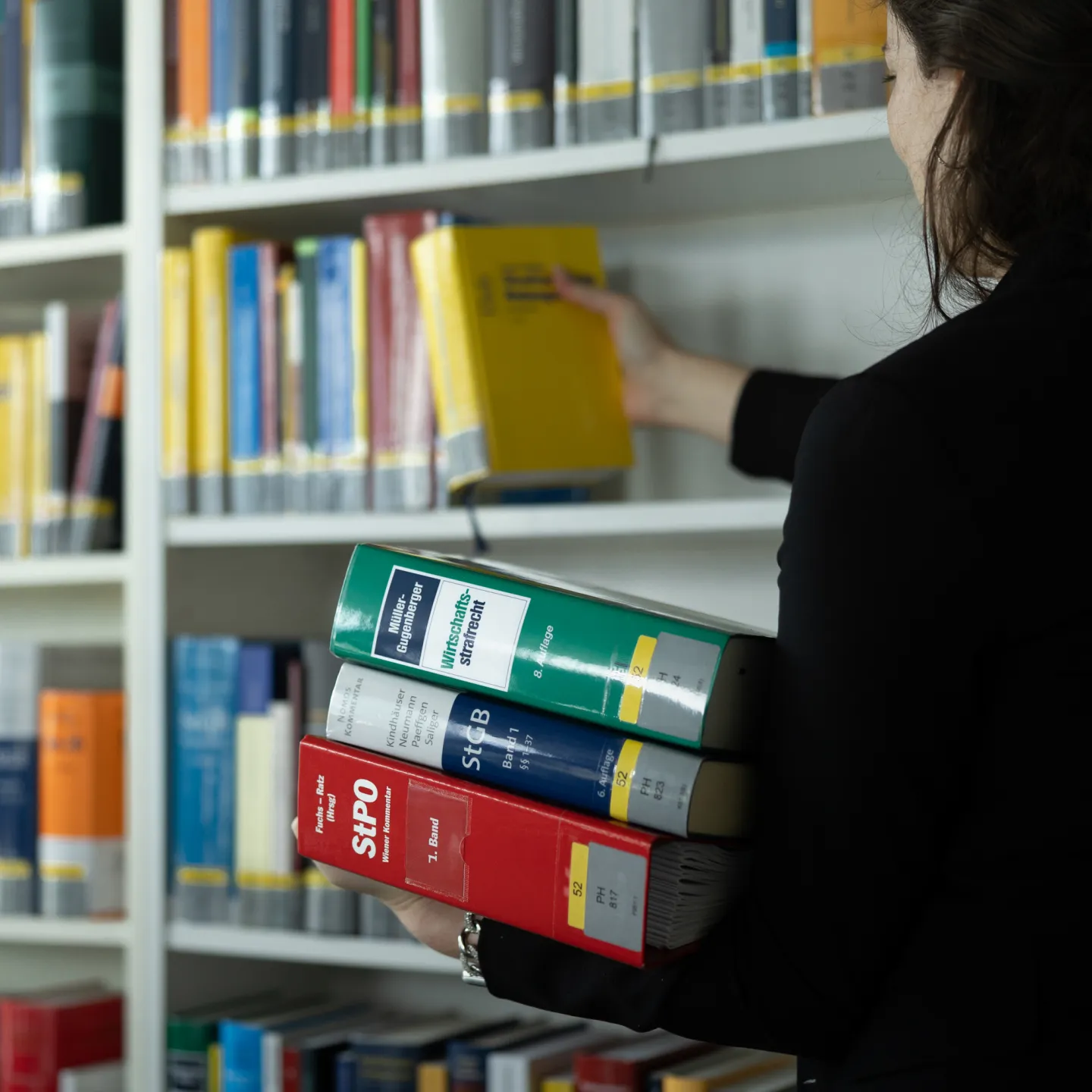 Frau stellt gelbes Fachbuch ins Regal und hält weitere juristische Bücher in der Bibliothek der Universität Liechtenstein.