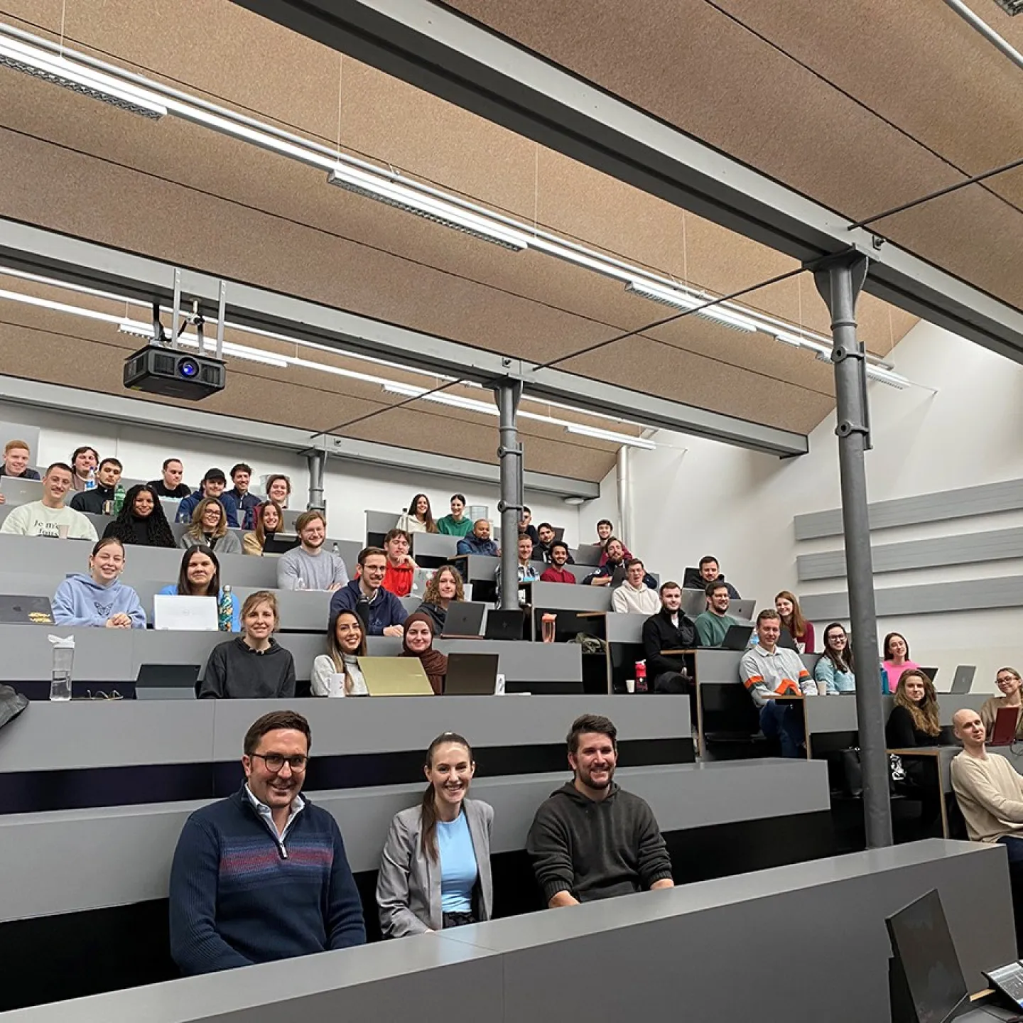 Eine Gruppe von Studierenden sitzt in einem modernen Hörsaal der Universität Liechtenstein. Sie nehmen an der Accenture Campus Innovation Challenge teil, einem Wettbewerb zur Entwicklung innovativer KI-Anwendungen. Die Studierenden lächeln und arbeiten an Laptops.