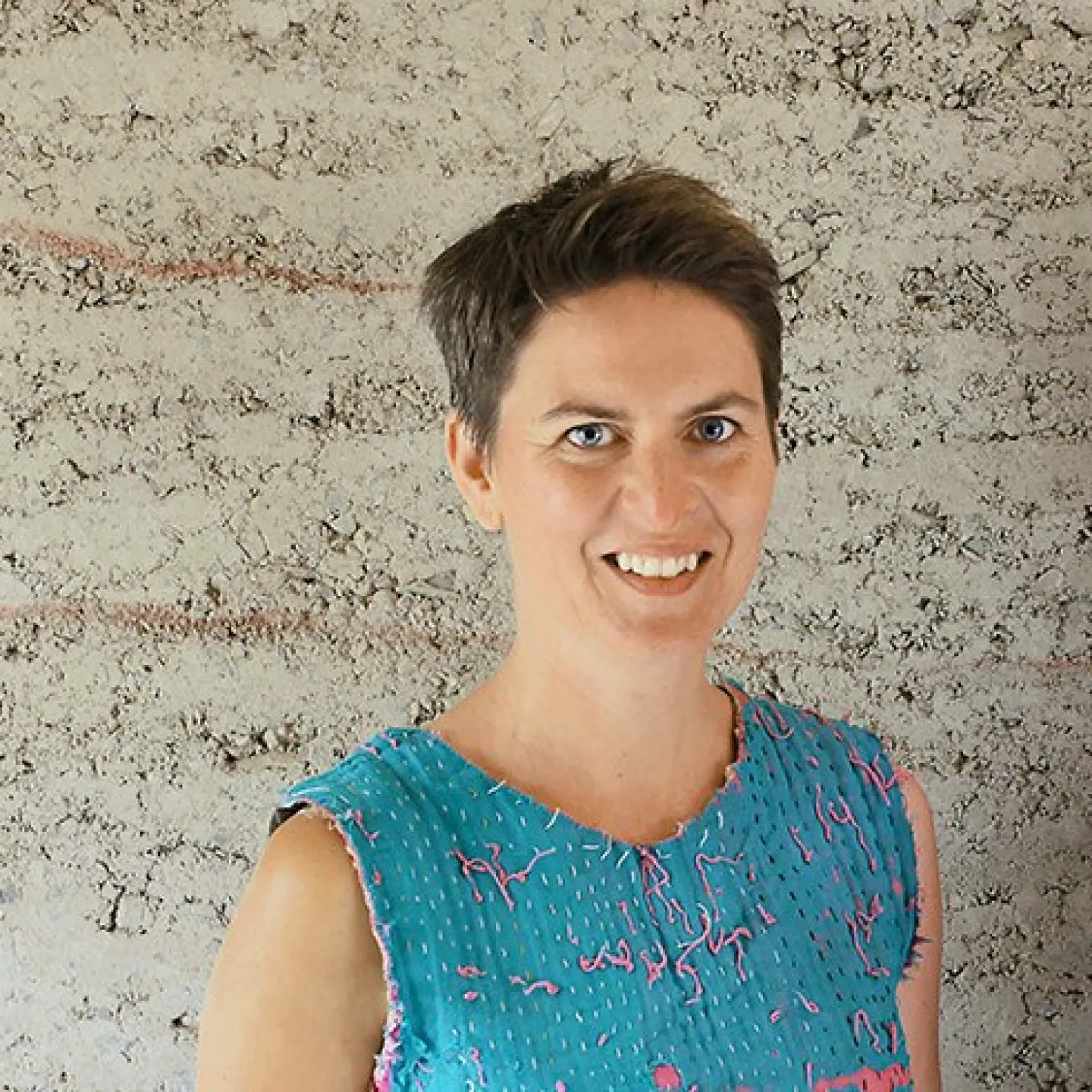 Eine lächelnde Person steht vor einer strukturierten Lehmwand. Sie trägt ein blaues Oberteil mit rosa Akzenten.