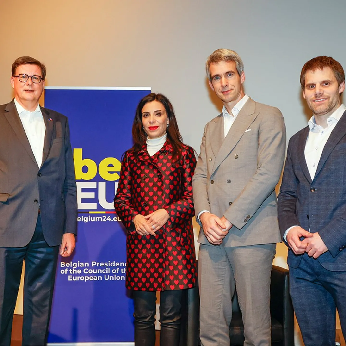 Vier Personen stehen nebeneinander vor einem Banner mit der Aufschrift "Belgian Presidency of the Council of the European Union". Sie tragen formelle Kleidung und blicken in die Kamera.
