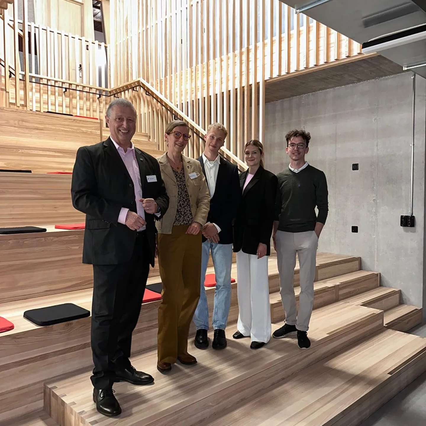 Fünf Personen stehen lächelnd in einer modernen Aula mit Holzstufen und vertikalen Holzlamellen im Hintergrund. Sie tragen formelle Kleidung und posieren für ein Gruppenfoto.