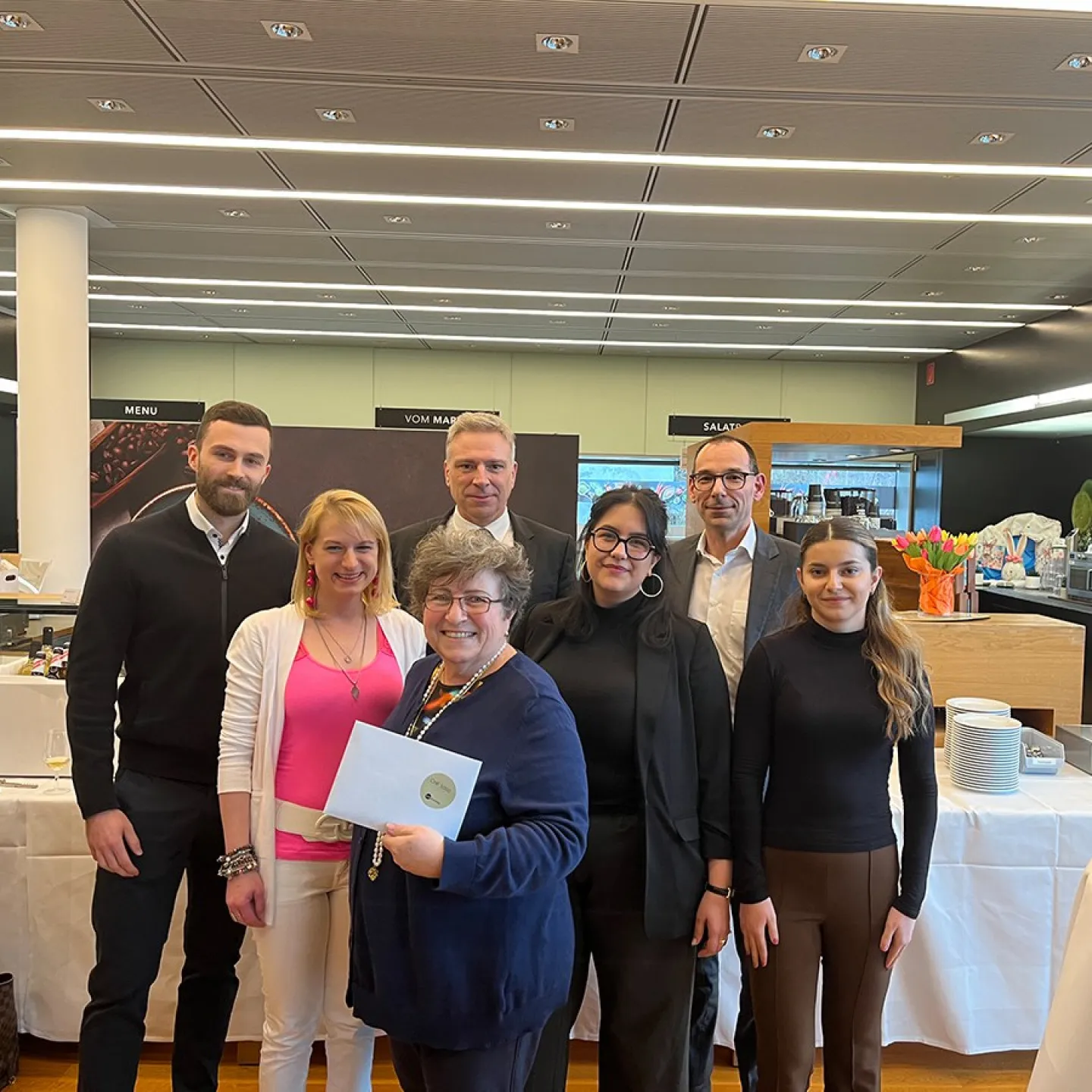 Eine Gruppe von sieben Personen steht lächelnd in einem modernen Raum. Eine Person hält ein Zertifikat in den Händen. Im Hintergrund sind Tische mit Geschirr und Dekorationen zu sehen.