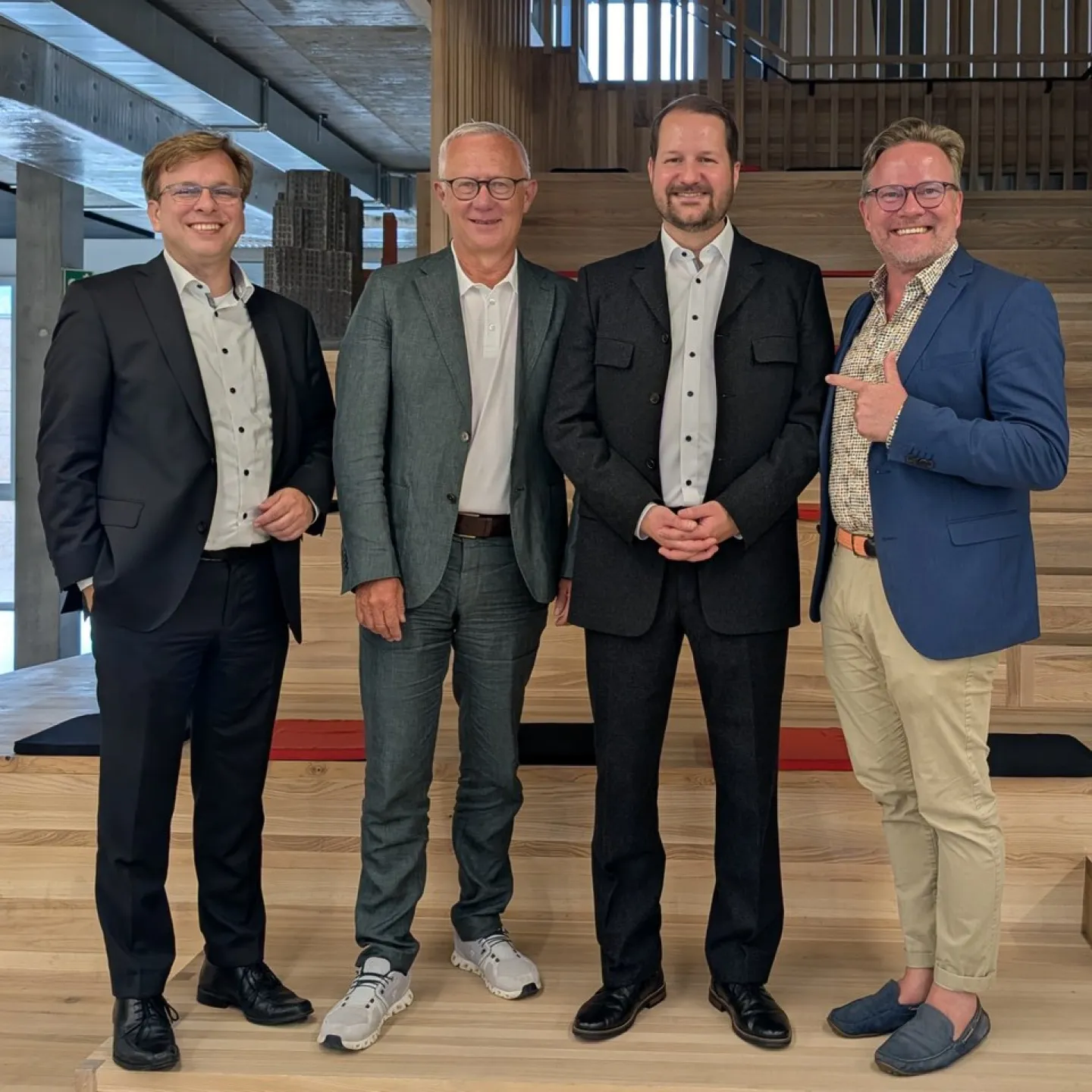 Vier Männer in formeller Kleidung stehen lächelnd auf einer Holztreppe in einem modernen Gebäude.